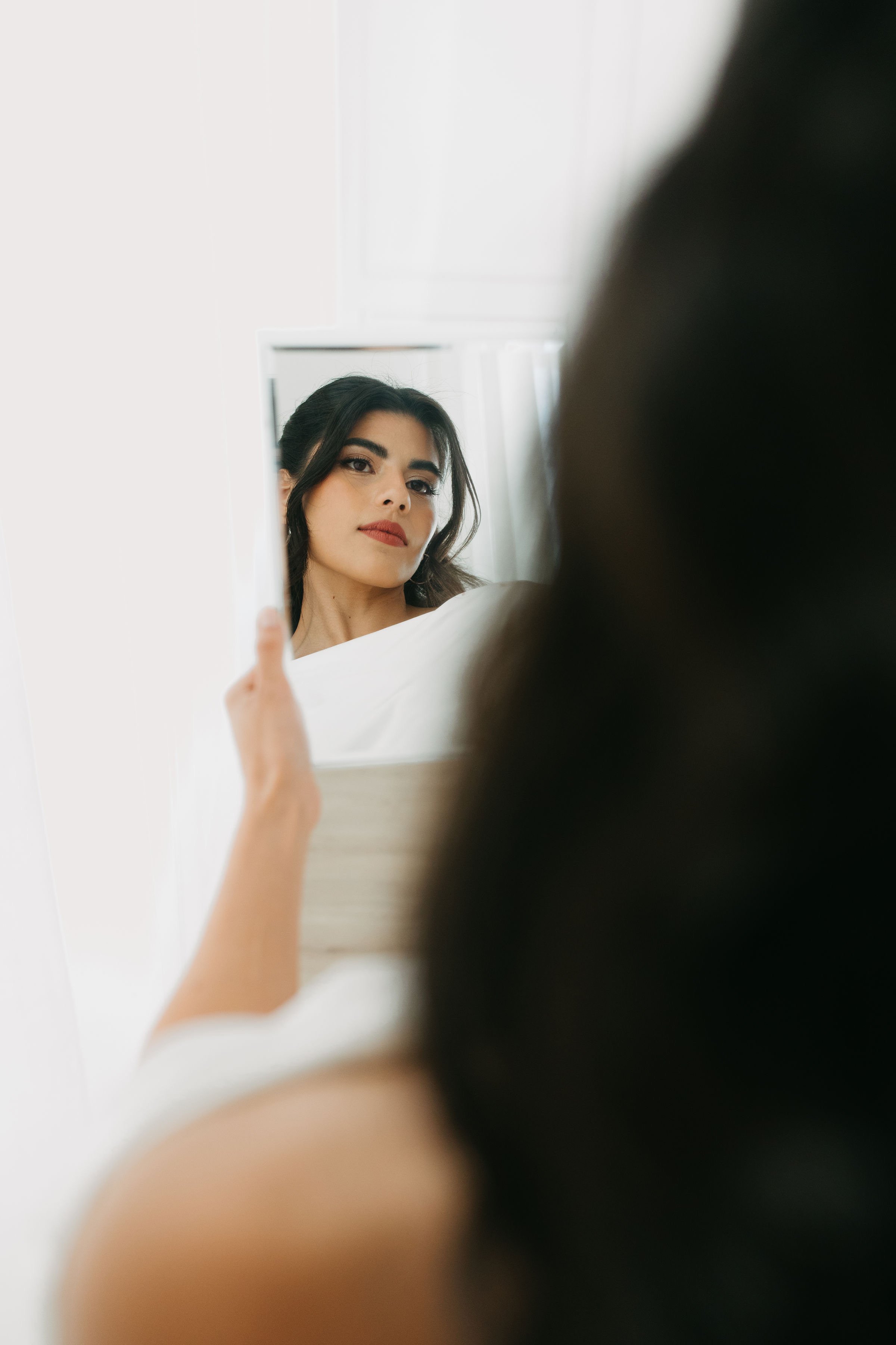 A woman with dark hair and makeup looking into a mirror, with the reflection showing her face.