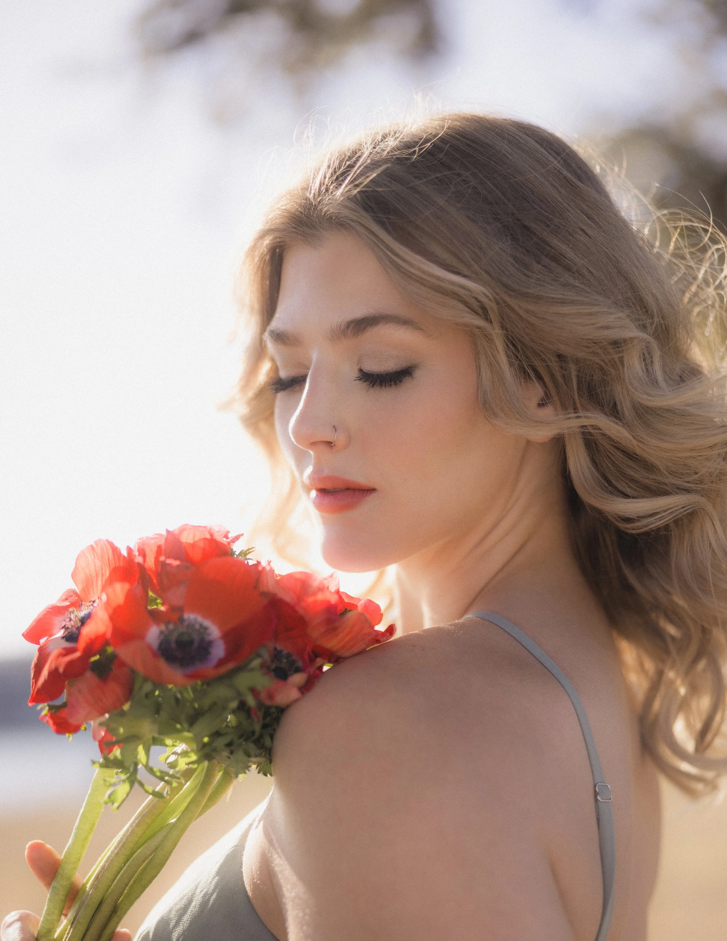A woman with blonde hair and a nose ring holding a bouquet of red and purple flowers with green stems. She is wearing a light gray strap of clothing and appears to be outdoors in sunlight.