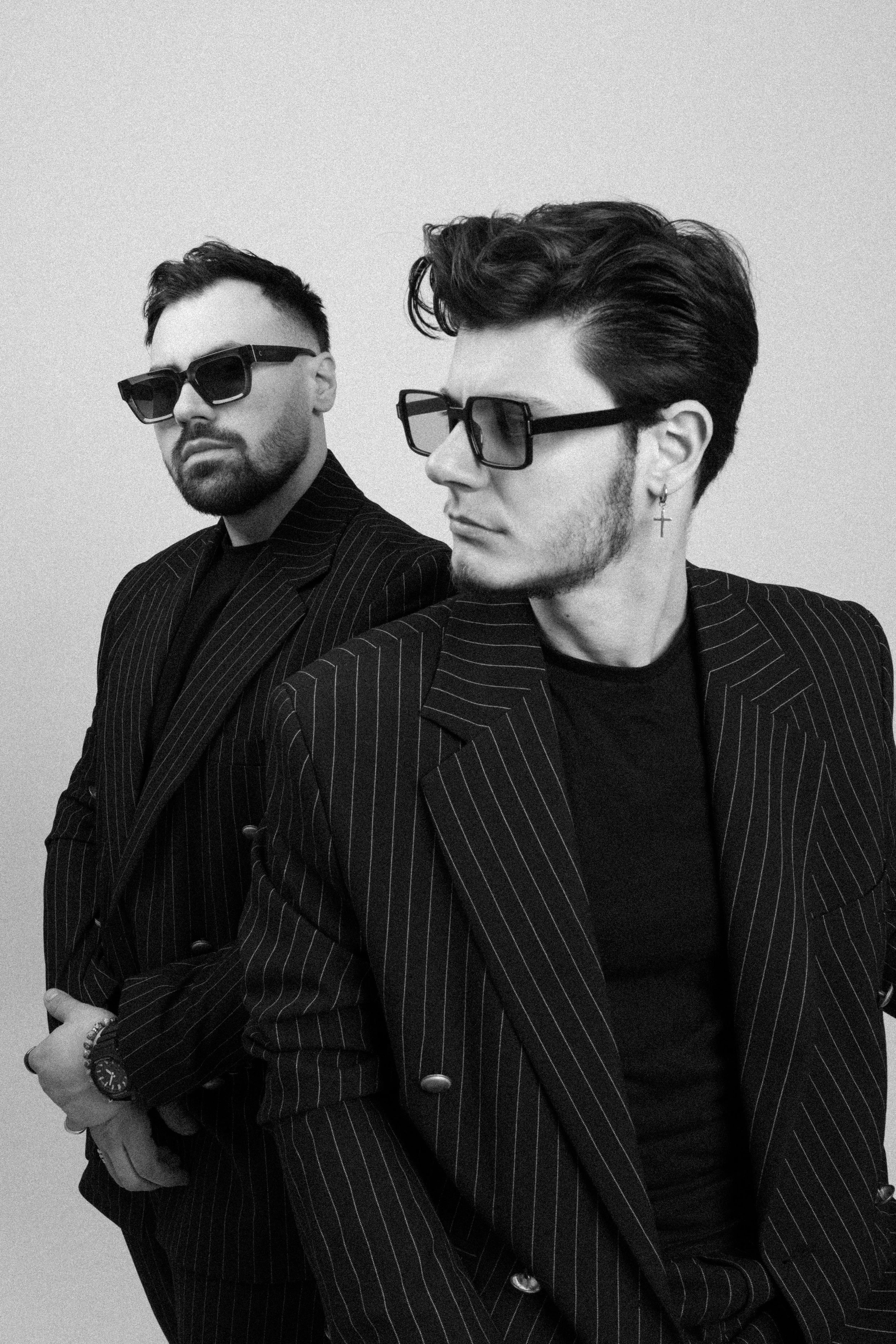 Two men with dark hair and beards, wearing black sunglasses and pinstripe suits, pose against a plain light background. One man has an earring and is turned sideways, while the other looks directly at the camera.
