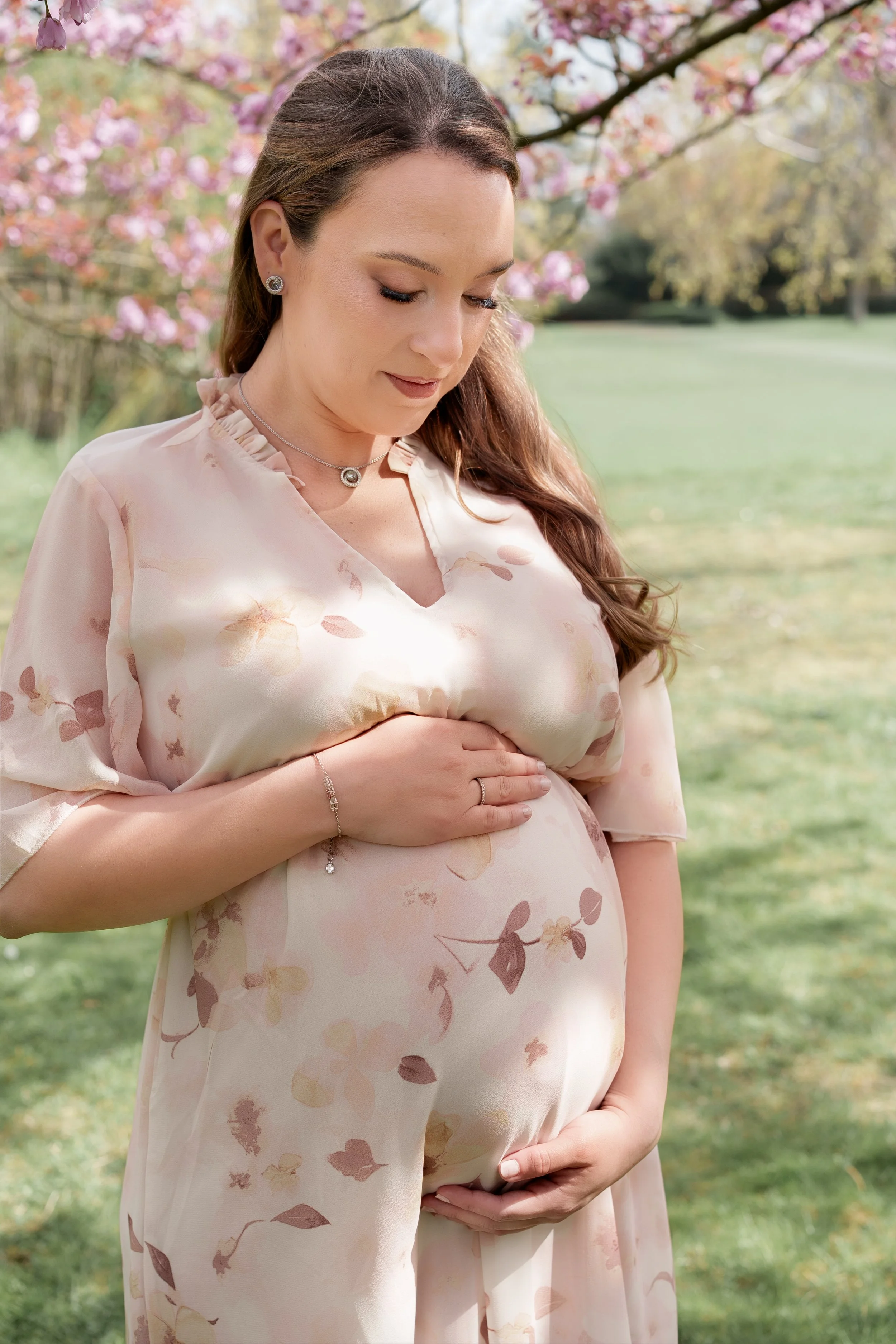 Eine schwangere Frau trägt ein helles, babyblaues Kleid mit rosa Blumenmuster und hält ihre Hände auf den Bauch. Sie steht im Freien mit blühenden Kirschbäumen im Hintergrund.