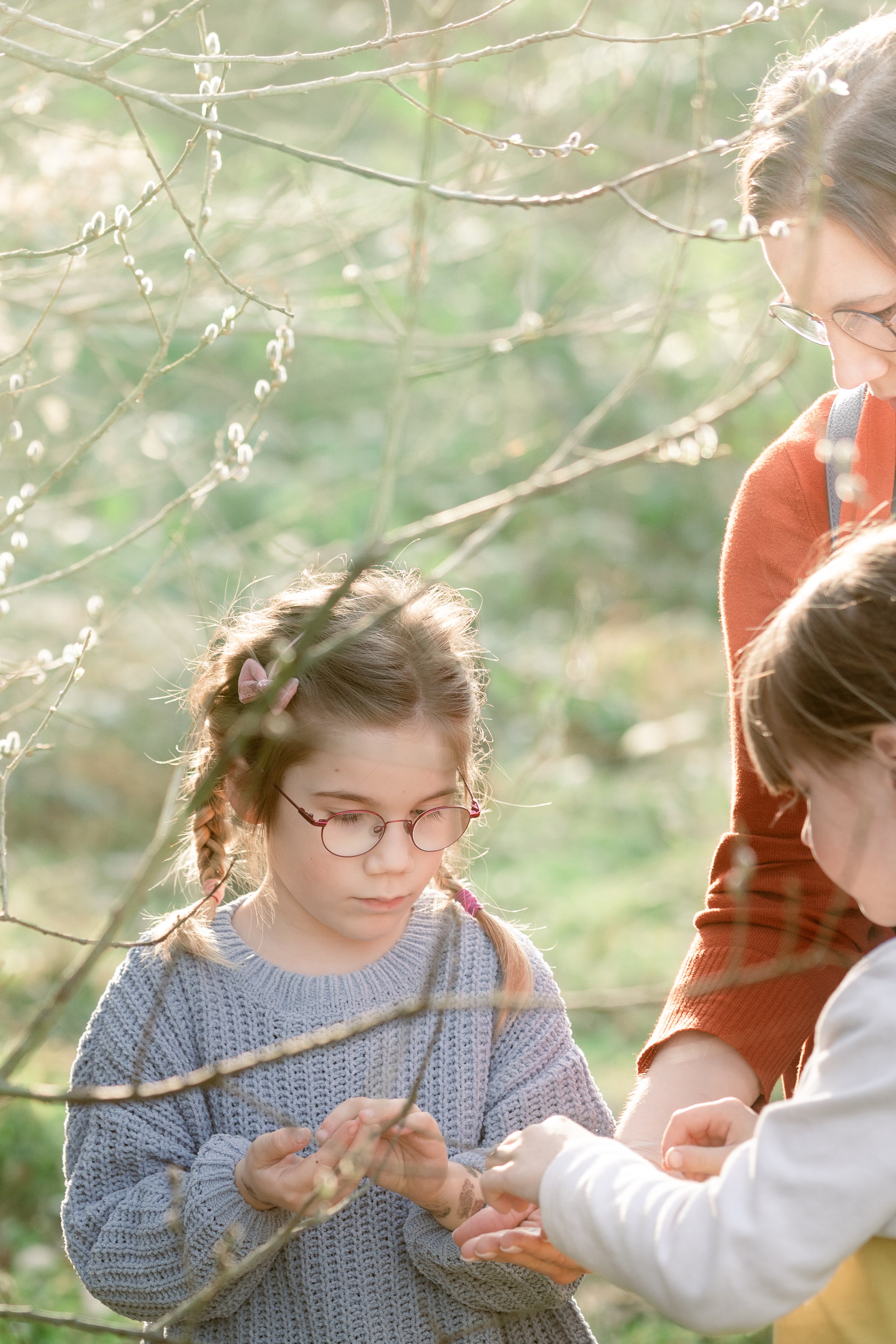 Eine junge Mädchen mit Brille und Zöpfen, die auf einem Ast in einem Wald steht, während eine Frau neben ihr ihr einen Gegenstand zeigt.