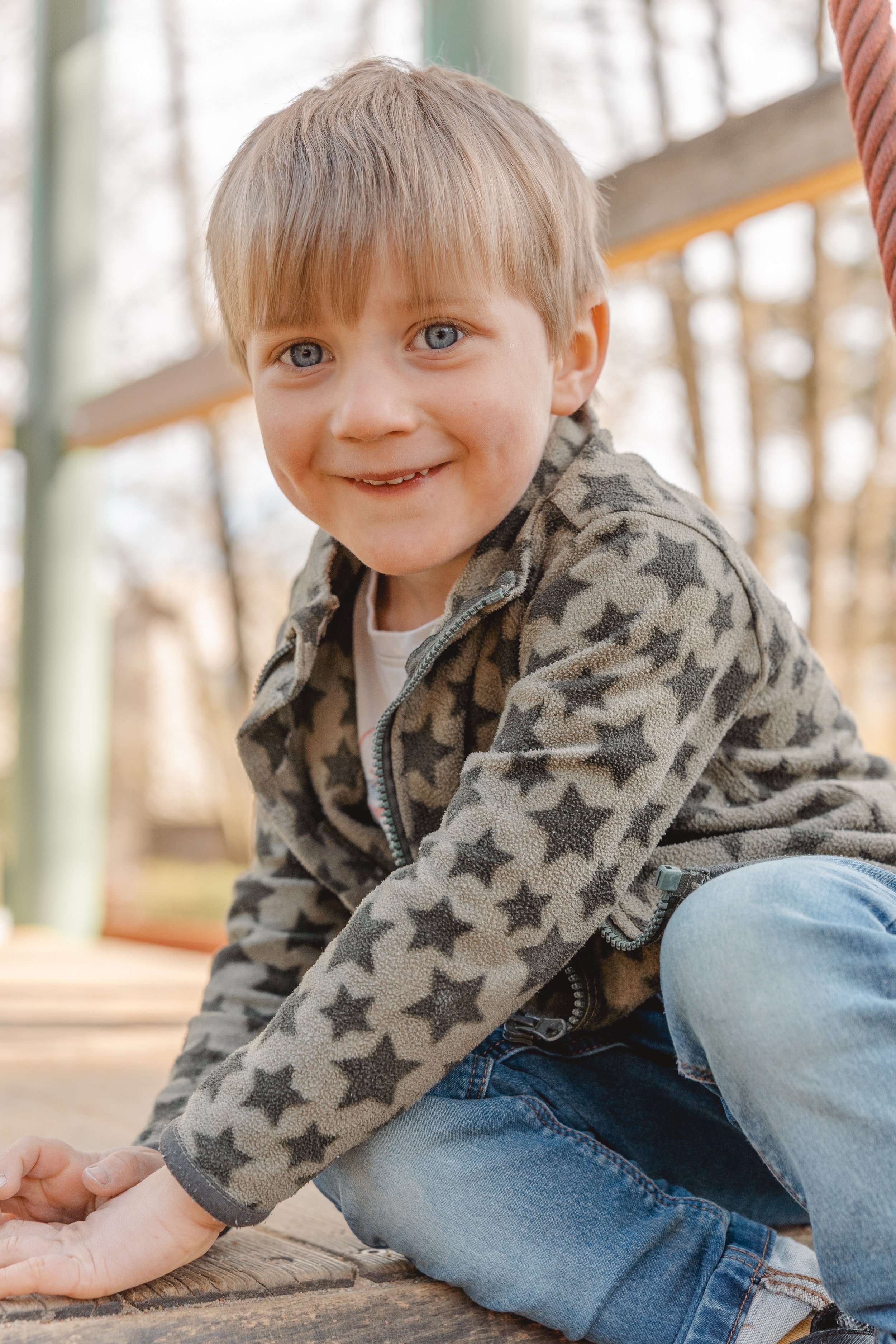 Junge mit blauen Augen und blonden Haaren, trägt einen Pullover mit Sternenmuster, spielt draußen auf einem Holzdeck, im Hintergrund Bäume.