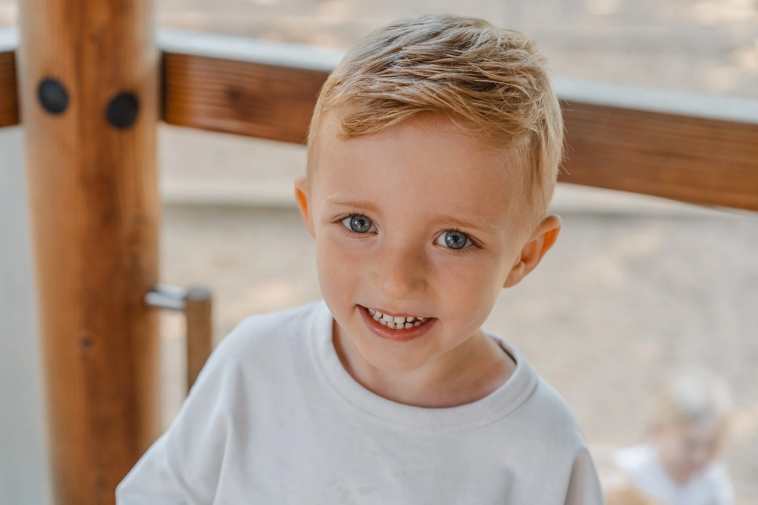 Ein lachender Junge mit blonden Haaren und blauen Augen, der ein weißes T-Shirt trägt, steht vor einem Spielgerät im Freien.