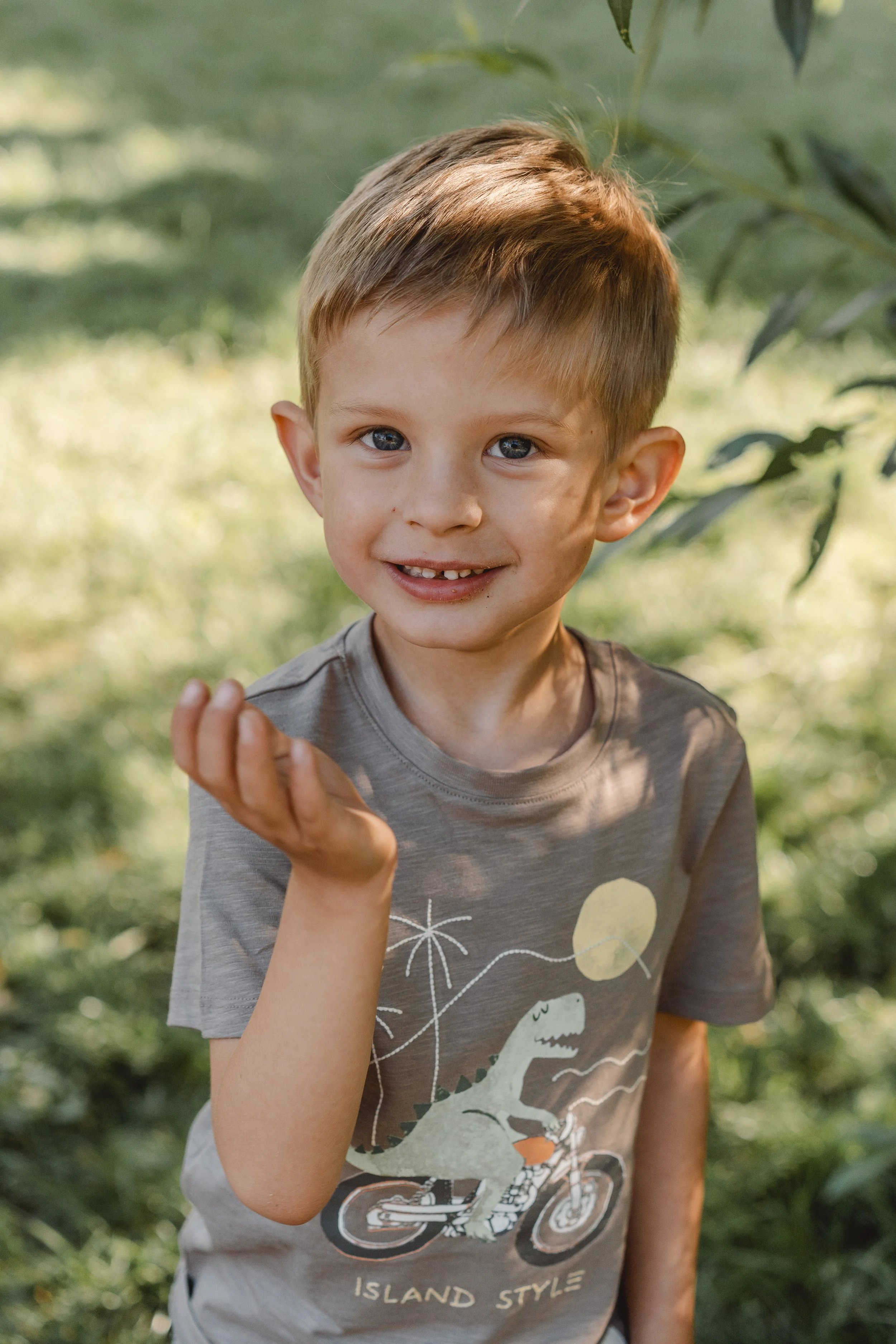 Ein lachender Junge mit blonden Haaren und blauen Augen trägt ein graues T-Shirt mit Dinosaurier-Designs im Freien, umgeben von grünem Laub.