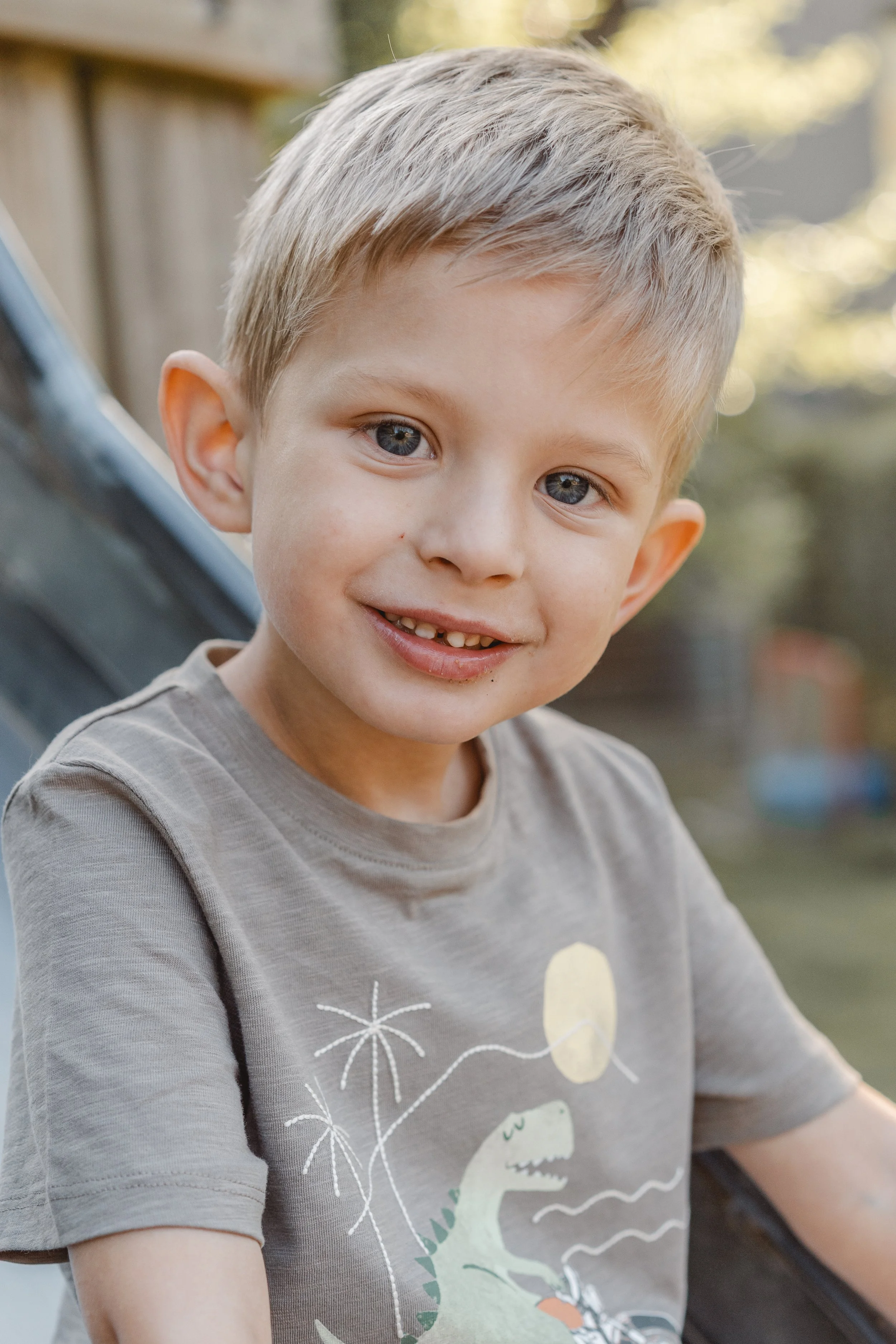 Ein Junge mit blonden Haaren und blauen Augen lächelt in die Kamera, trägt ein graues T-Shirt mit Dinosaurier-Design, im Freien bei Sonnenlicht.