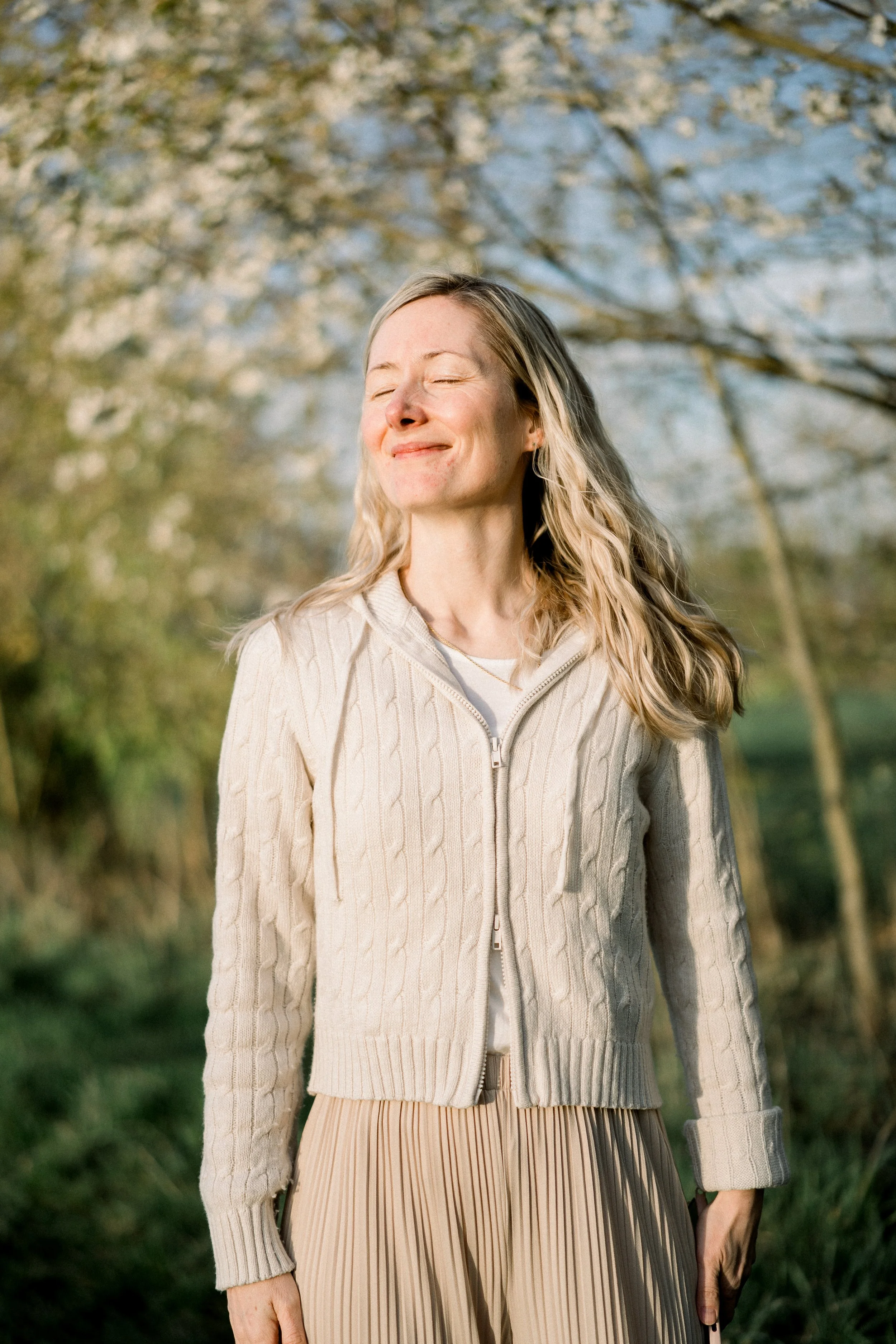 Eine lachende Frau mit geschlossenen Augen steht im Freien an einem sonnigen Tag und genießt die Sonne, im Hintergrund sind Bäume mit Blättern.