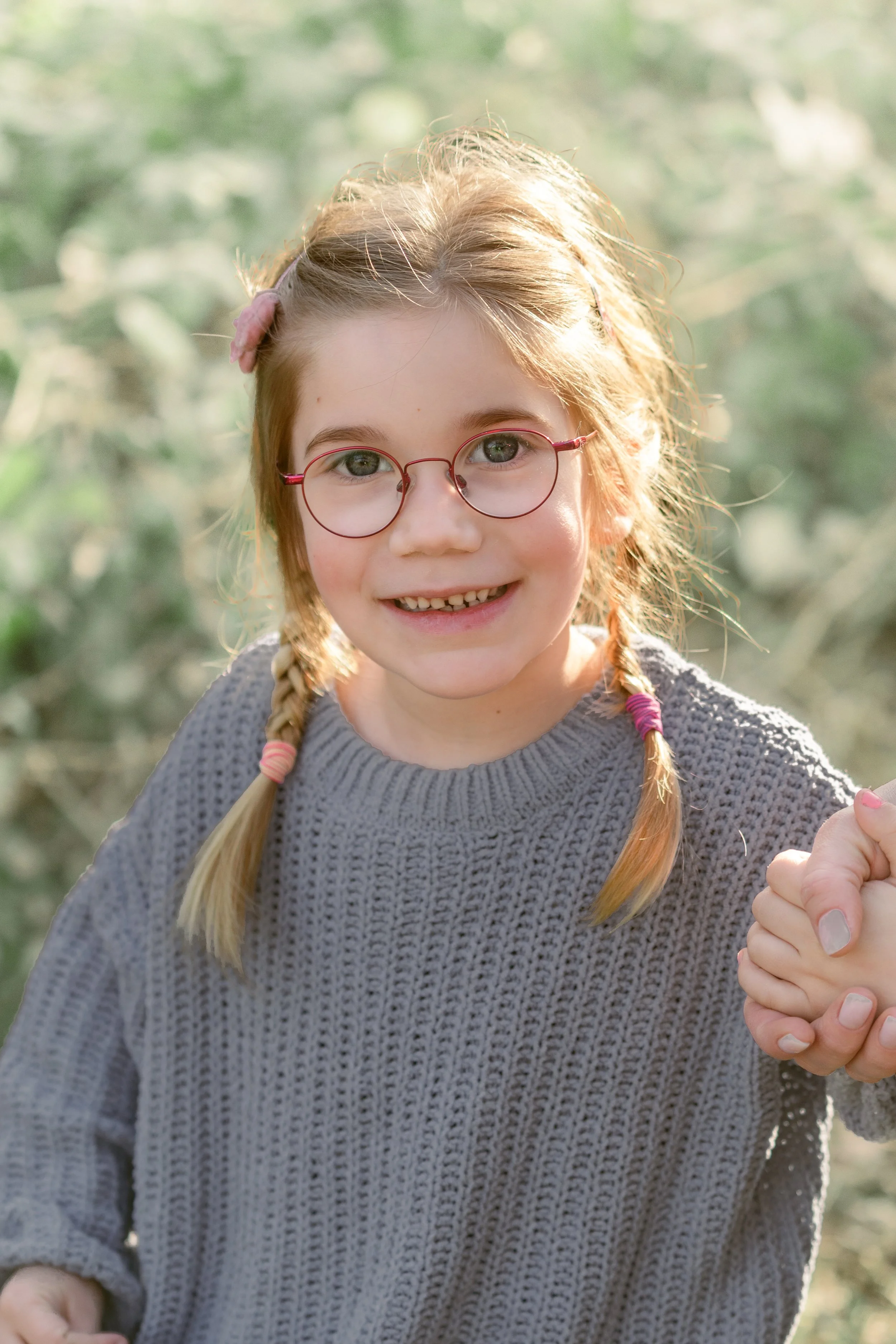 Ein lachendes Mädchen mit Brille, Zöpfen und einem grauen Pullover, das draußen im Sonnenschein ist.