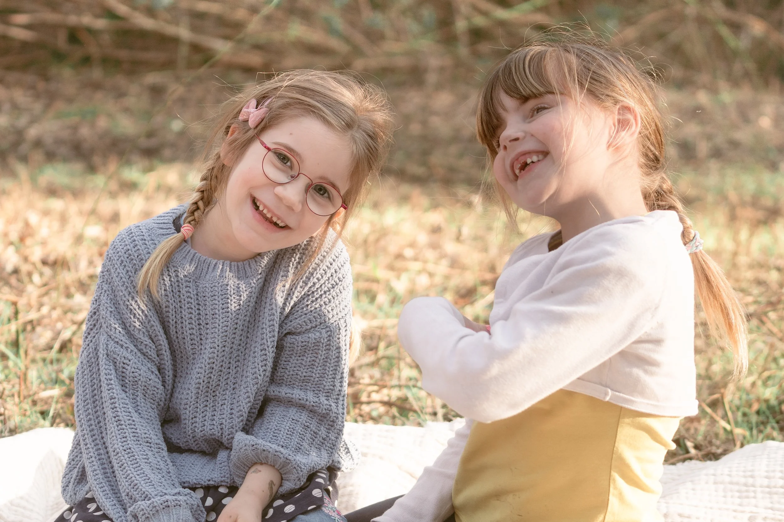 Zwei lachende junge Mädchen sitzen im Freien auf einer Decke im Wald, eines trägt eine graue Strickjacke und Brille, das andere ein weißes Oberteil mit gelbem Kleid darunter.