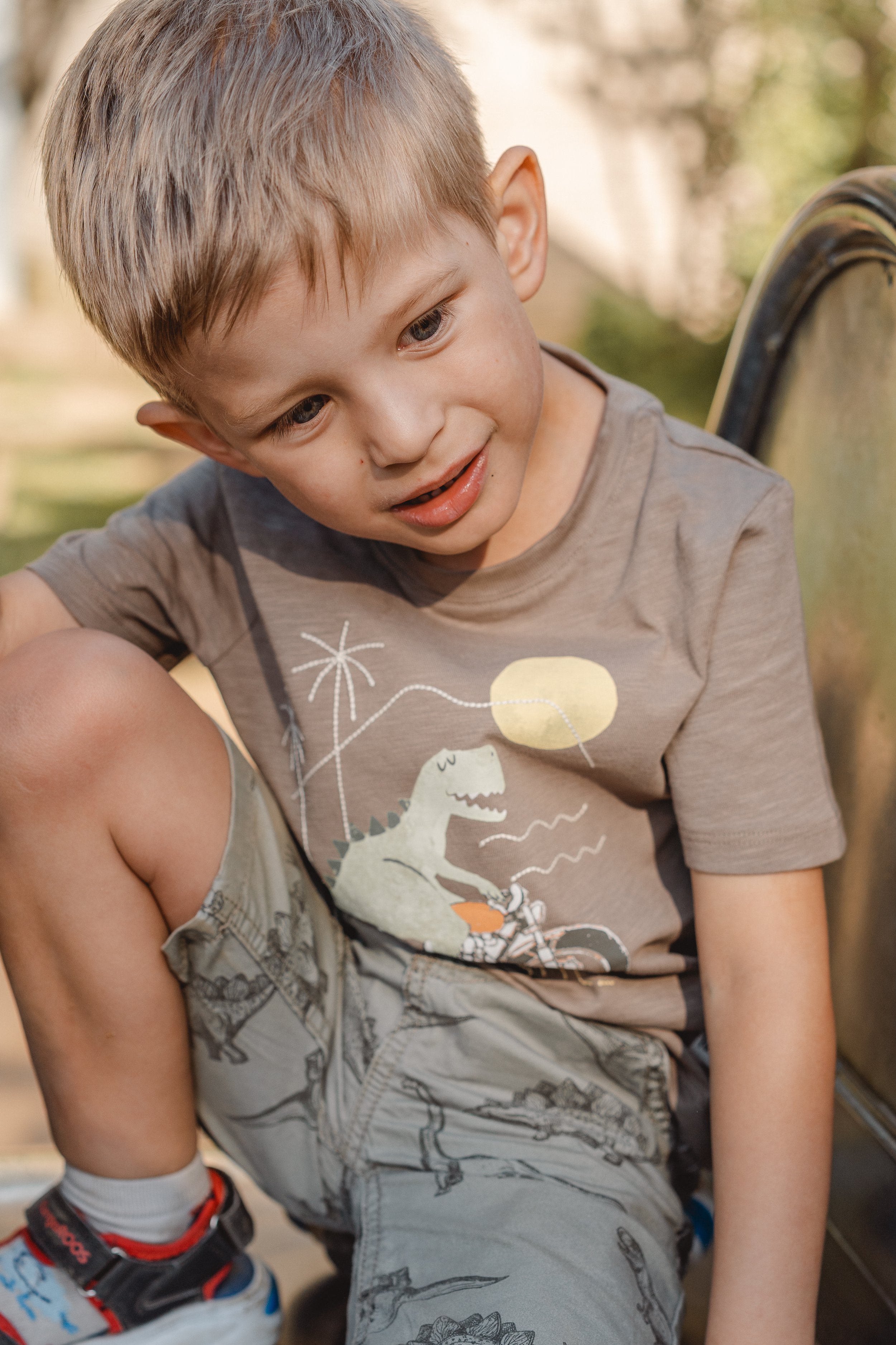 Ein junger Junge mit blonden Haaren sitzt draußen auf einem Stuhl, trägt ein graues T-Shirt mit Dinosaurier- und Ballmotiven und schaut nach unten. Im Hintergrund sind Bäume und Sonnenlicht sichtbar.