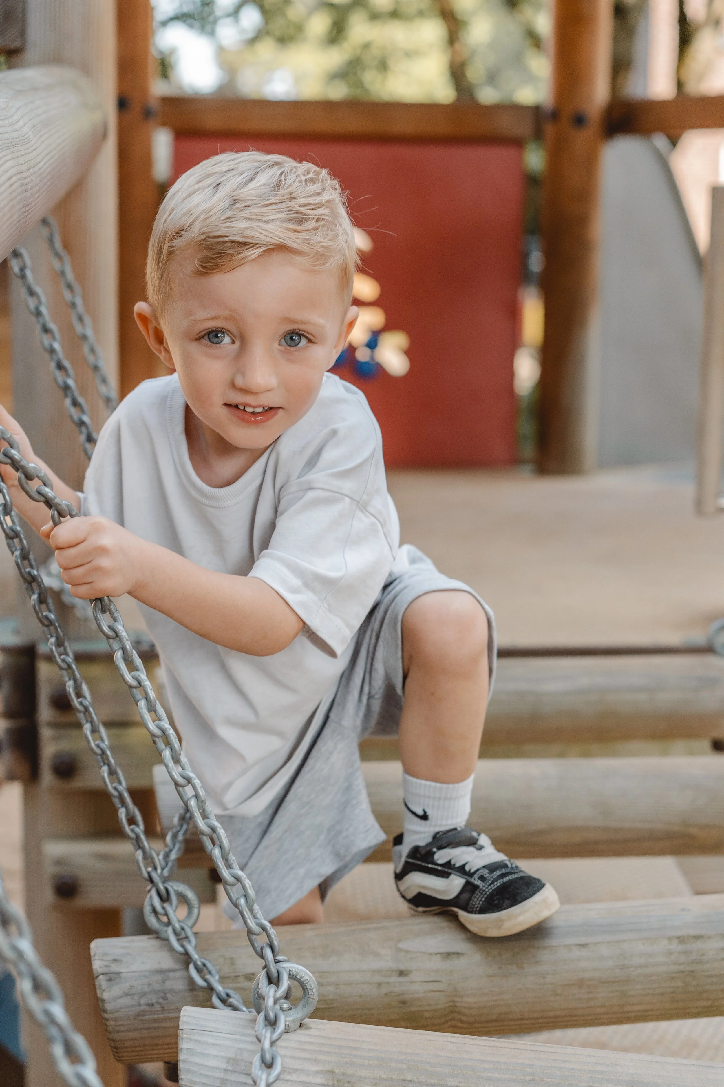 Ein junger Junge mit blonden Haaren, blauen Augen und einem weißen T-Shirt, der auf einem Spielplatz auf den Holzboden vor einem Kletternetz sitzt und mit den Händen nach oben greift, um sich festzuhalten. Im Hintergrund sind Baumstämme und eine rote Wand des Spielgerüsts sichtbar.