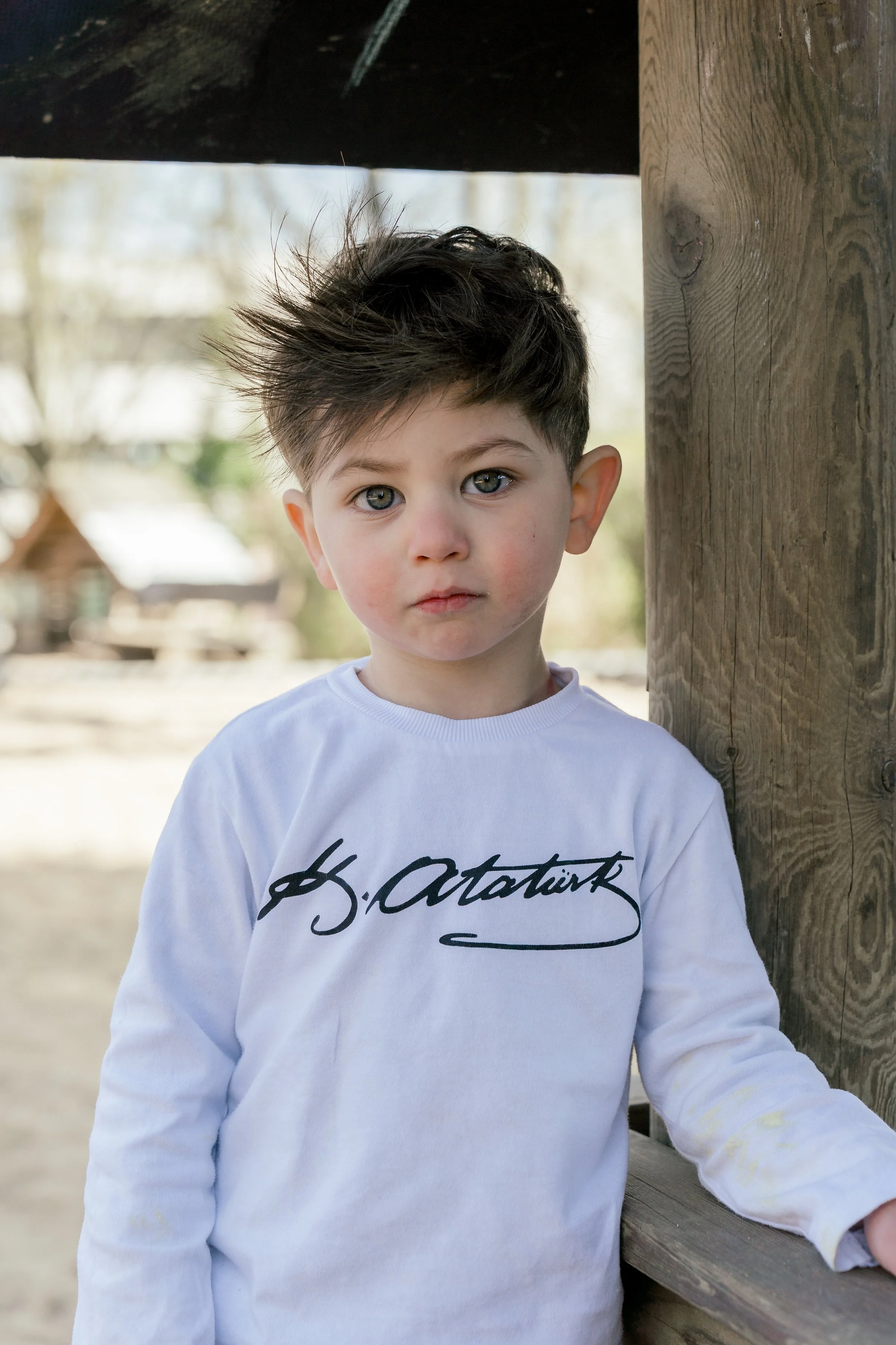 Junge mit dunklem, zerzaustem Haar, trägt weißes Sweatshirt, steht an Holzkonstruktion im Freien, unscharfer Hintergrund mit Bäumen und Gebäuden.