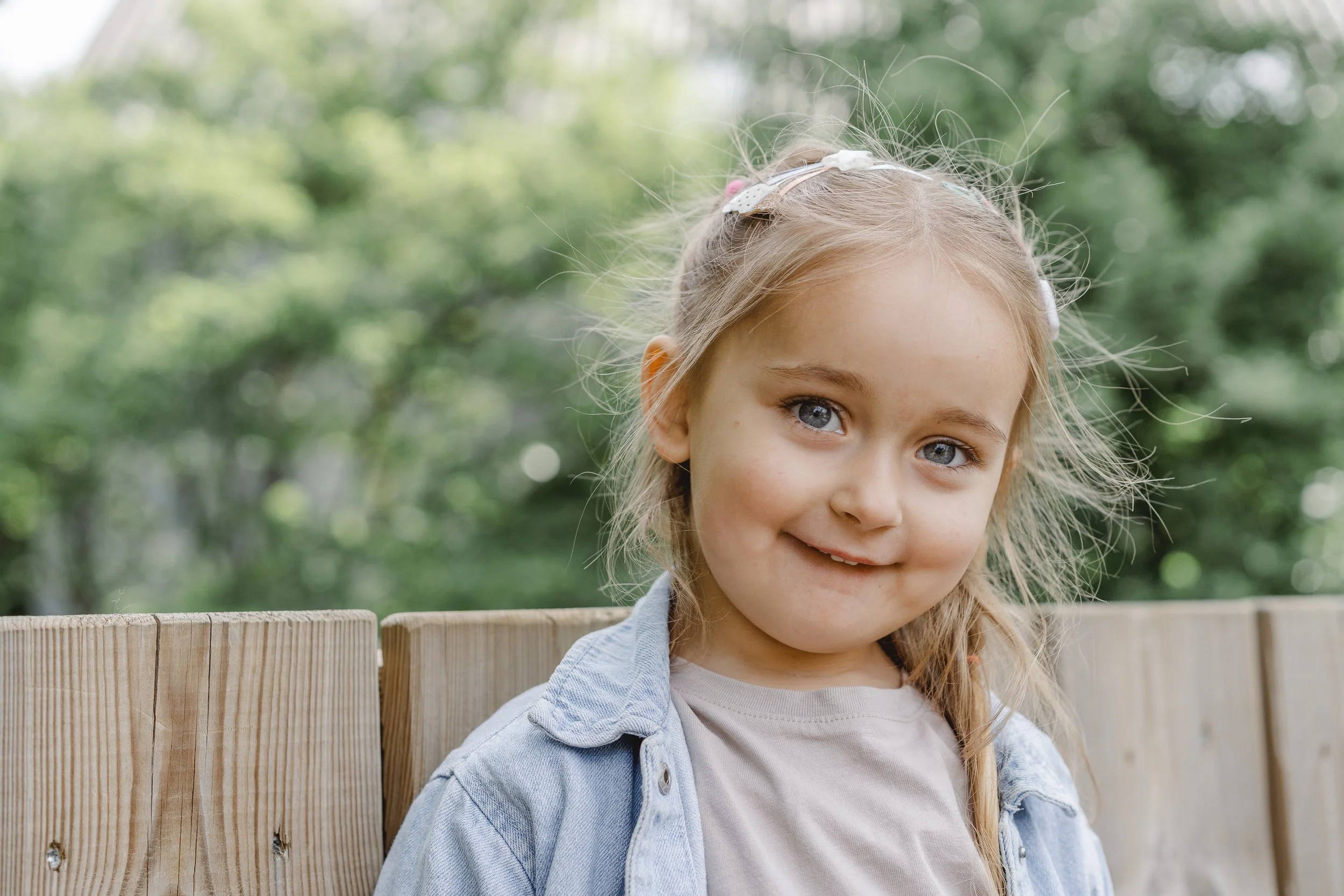 Nahaufnahme eines lachenden Mädchens mit blonden Haaren und blauen Augen, das im Freien sitzt, mit einem Holzzaun im Vordergrund und grünem, unscharfem Hintergrund.