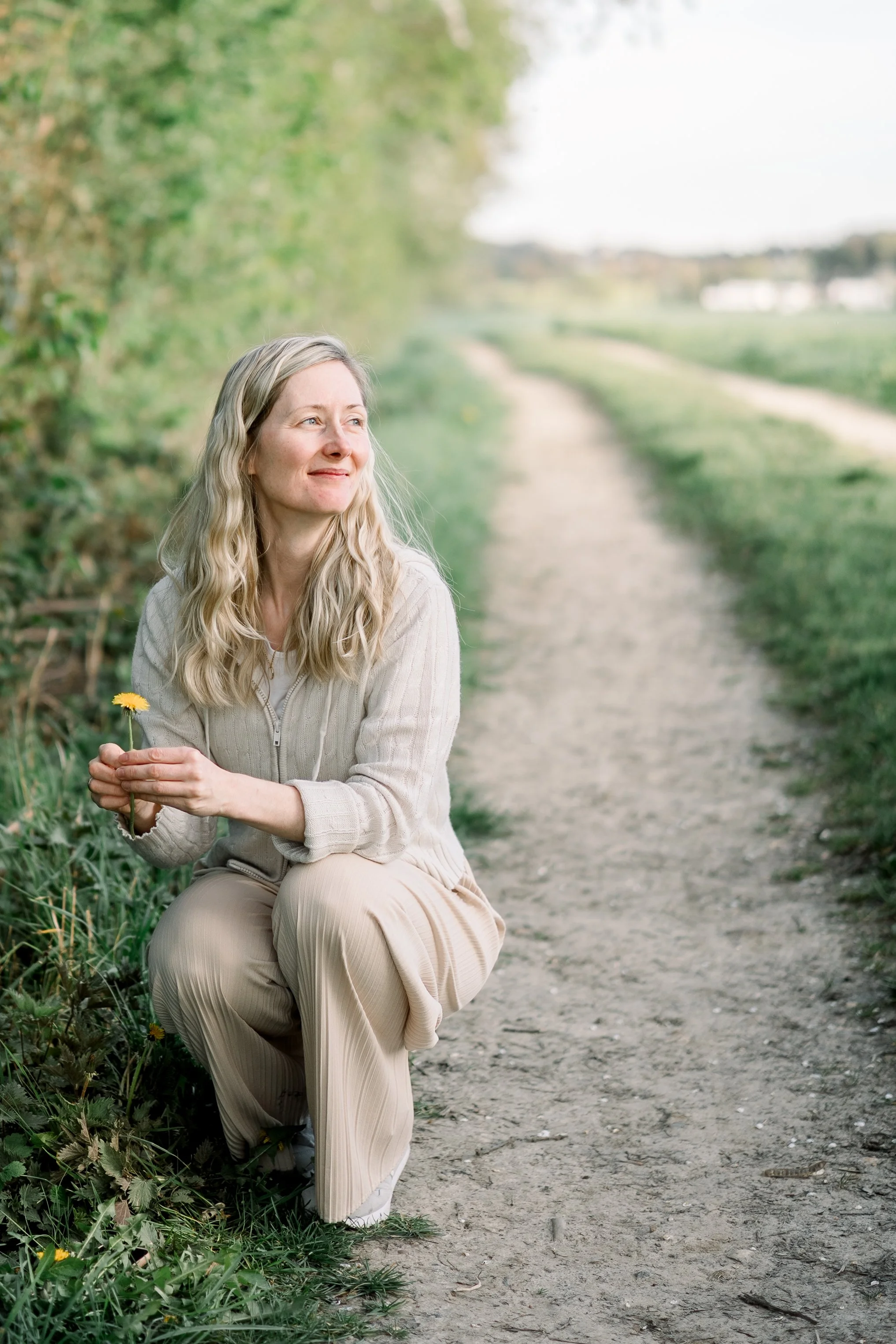 Eine Frau mit blonden, welligen Haaren, die neben einem Feldweg im Freien sitzt, ein gelber Gänseblümchen in der Hand, mit einem entspannten und glücklichen Gesichtsausdruck.