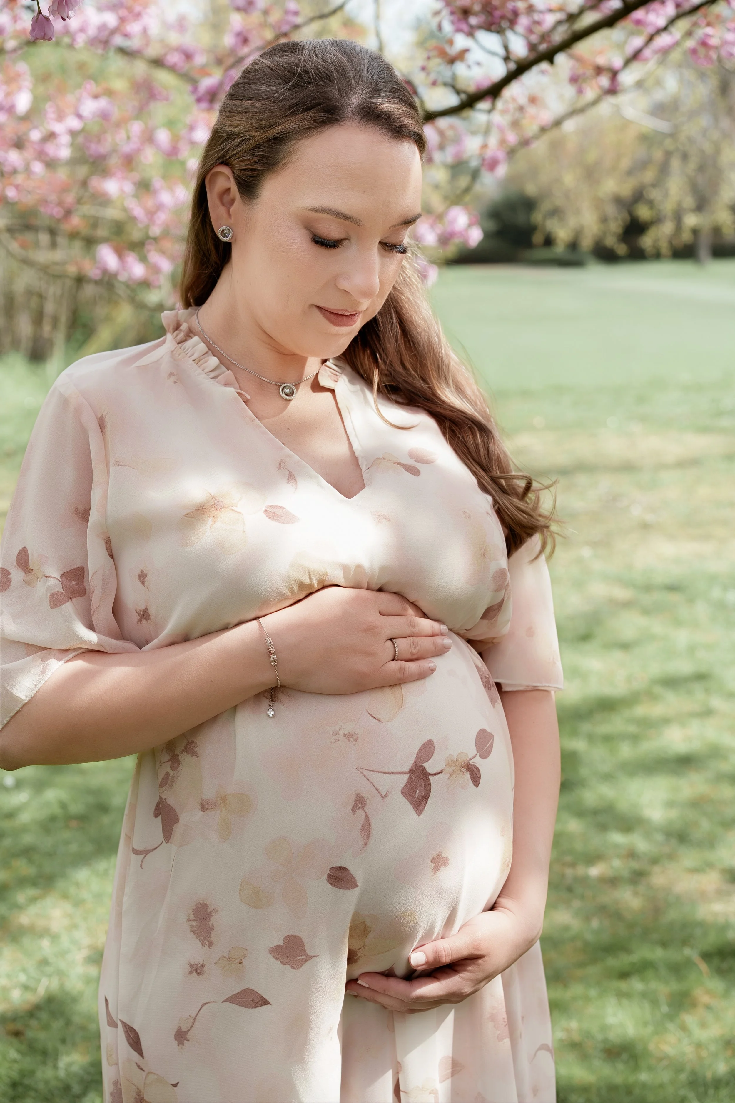 Schwangere Frau in einem hellen Kleid mit Blumenmuster, die den Bauch streichelt, im Park mit blühenden Bäumen im Hintergrund.