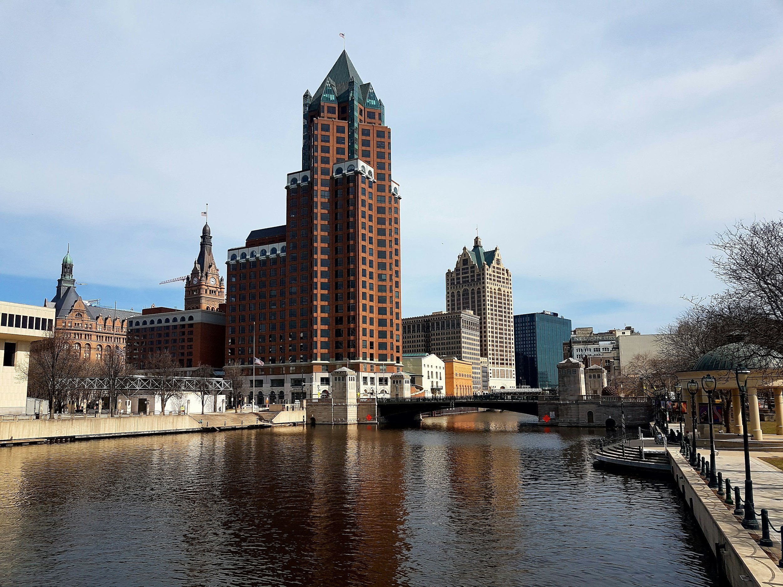 City skyline featuring tall buildings along a river, with a bridge and a walkway with lamps and trees on the right side.