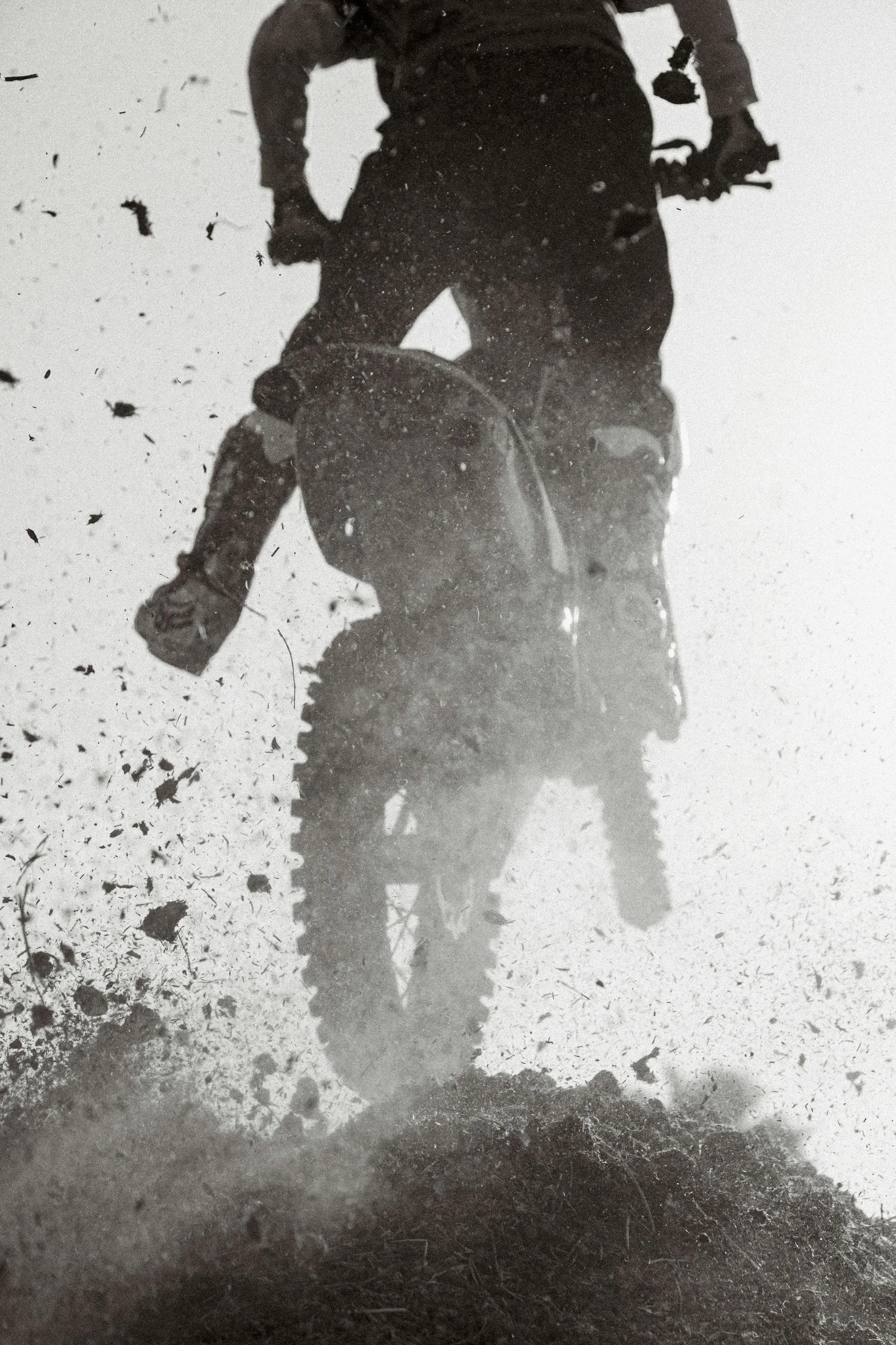 A person riding a dirt bike on a dirt trail, with dirt and small rocks kicking up around the bike. The rider is wearing protective gear and is captured from a low angle.