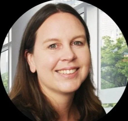 A woman with long brown hair smiling, with a background of modern office windows and green trees outside.