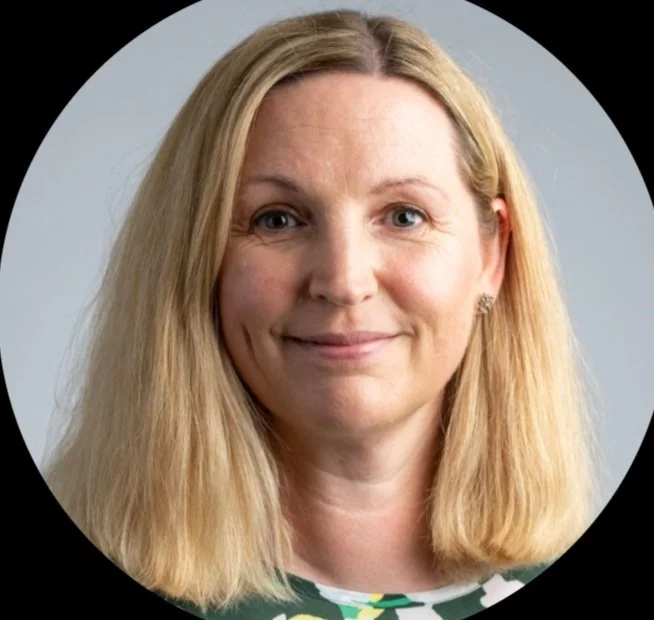 Portrait of a woman with blonde hair, smiling, wearing a green patterned top, against a light gray background.