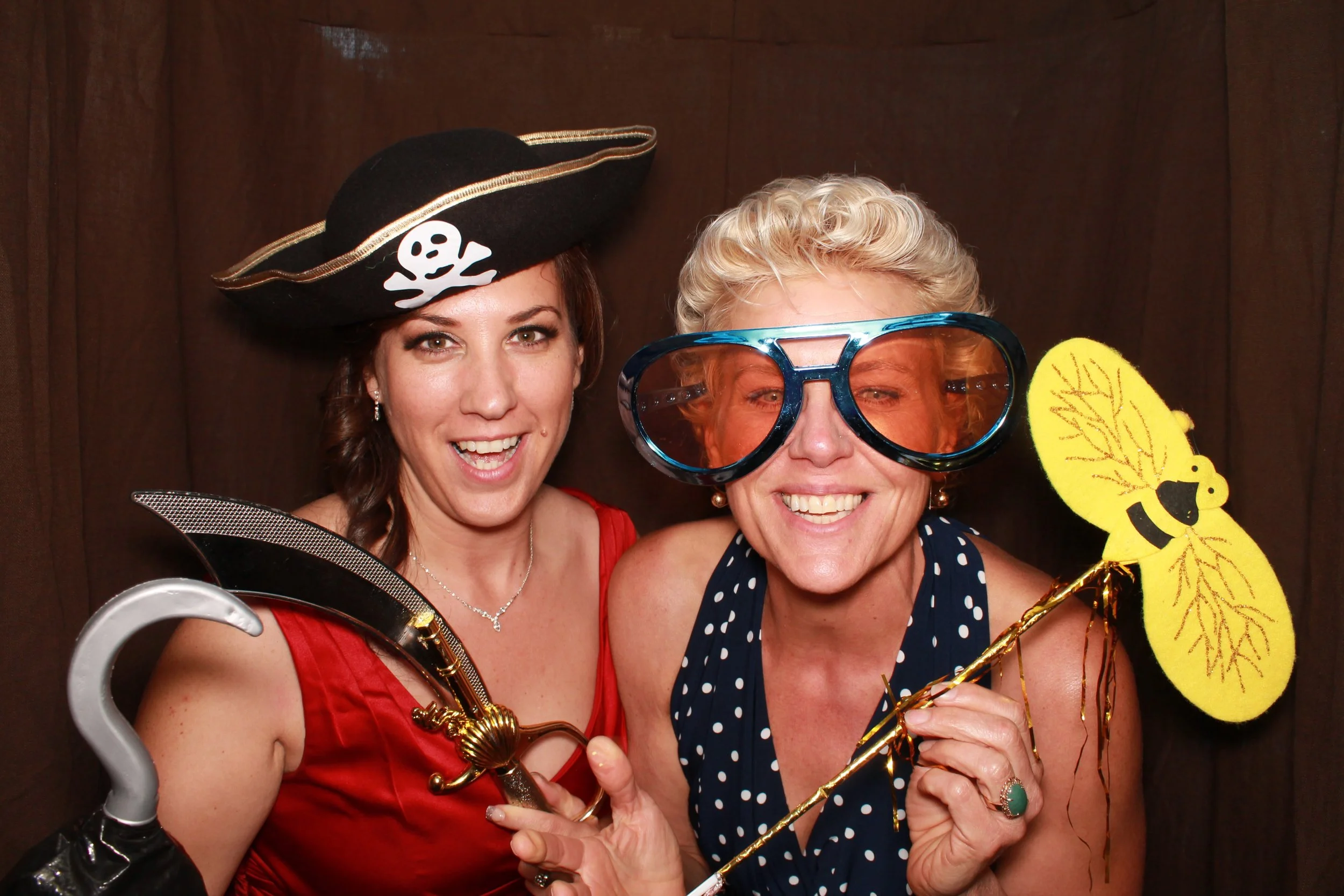  Two women smiling in an event photo booth with props in Burnaby, one donning a pirate hat, the other with oversized glasses and a bee prop. 