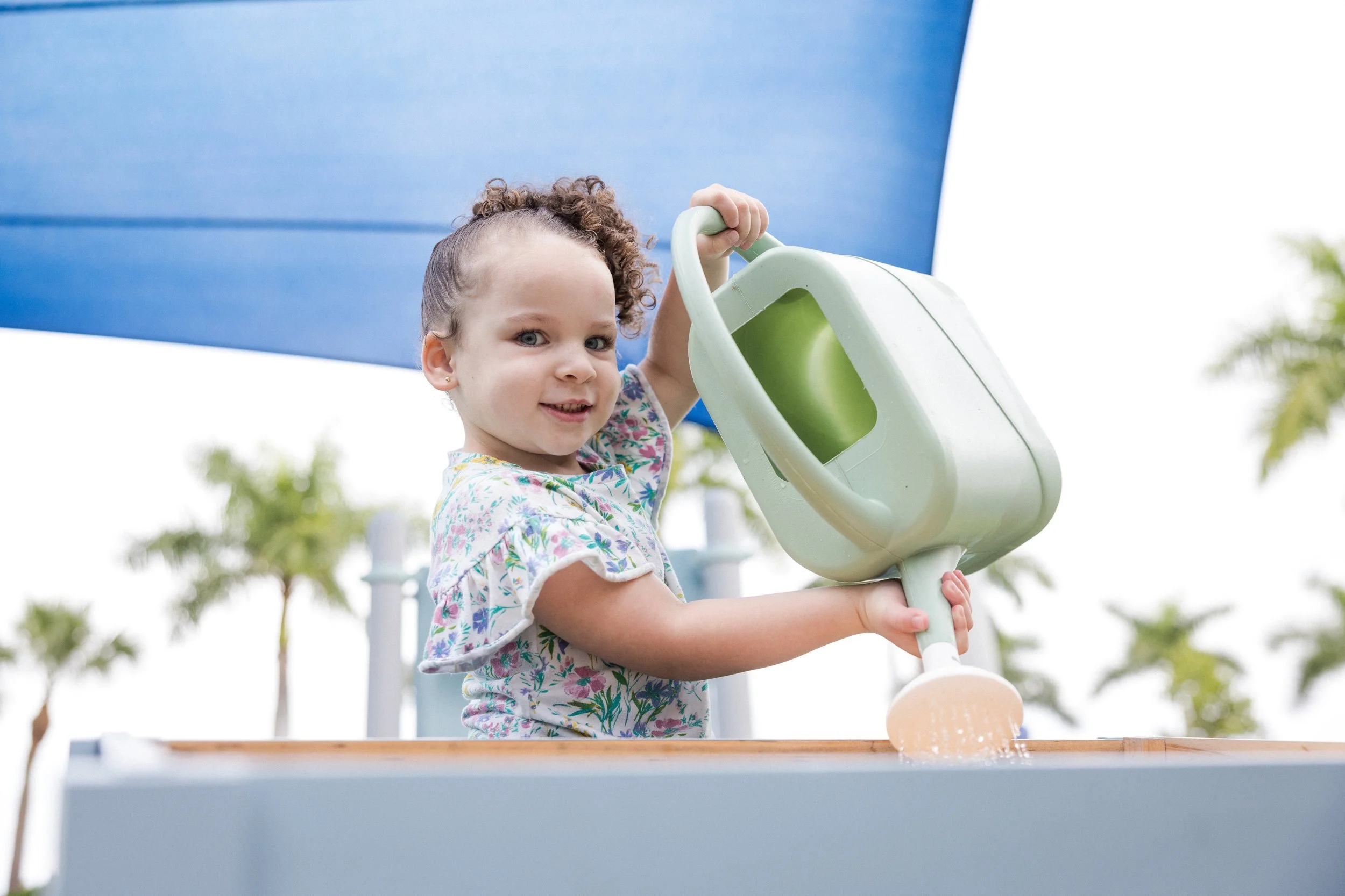 A young girl playing outdoors, pouring water from a green watering can over a wooden surface.