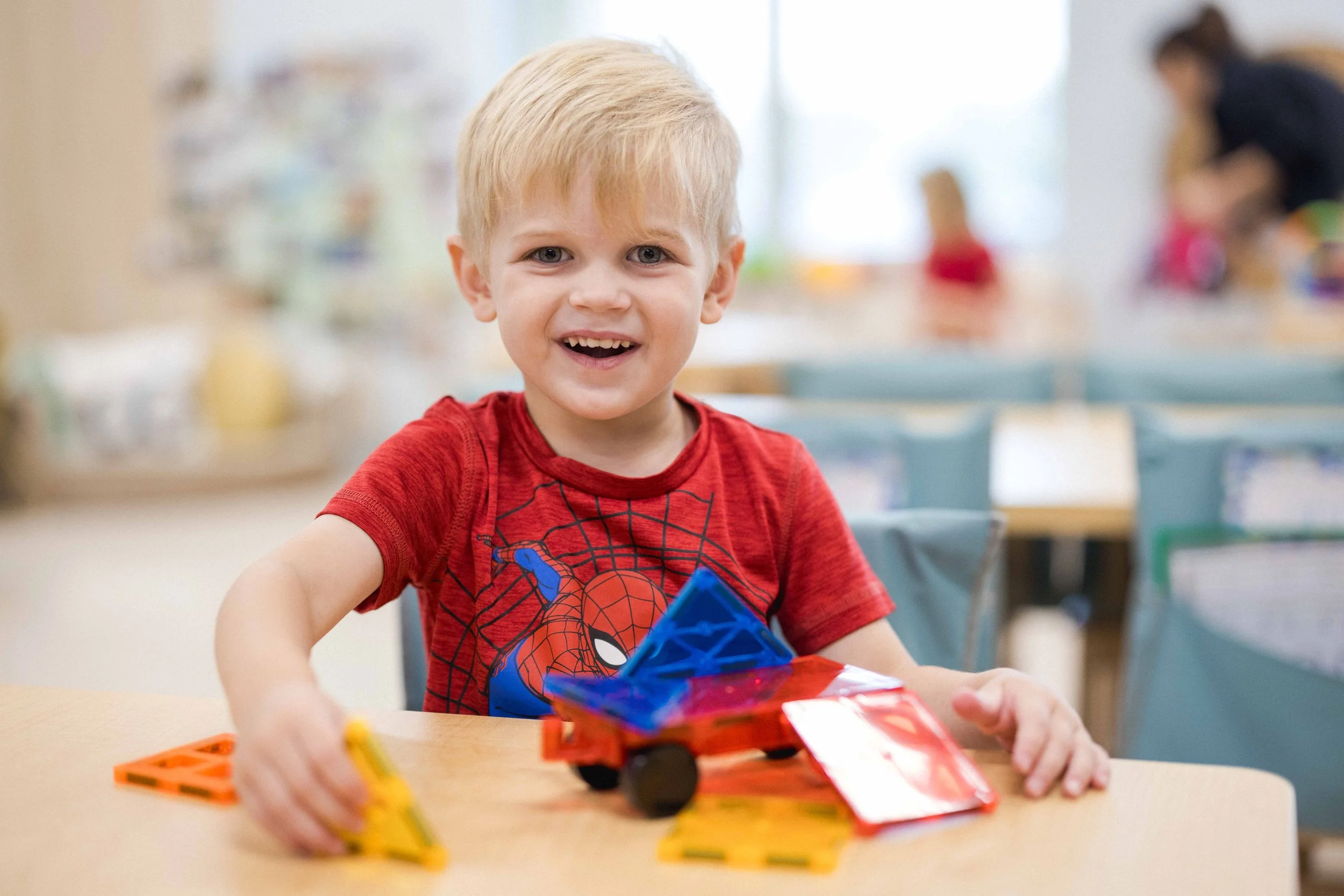 A young boy with blonde hair smiling and playing with magnetic construction toys at a table in a classroom.
