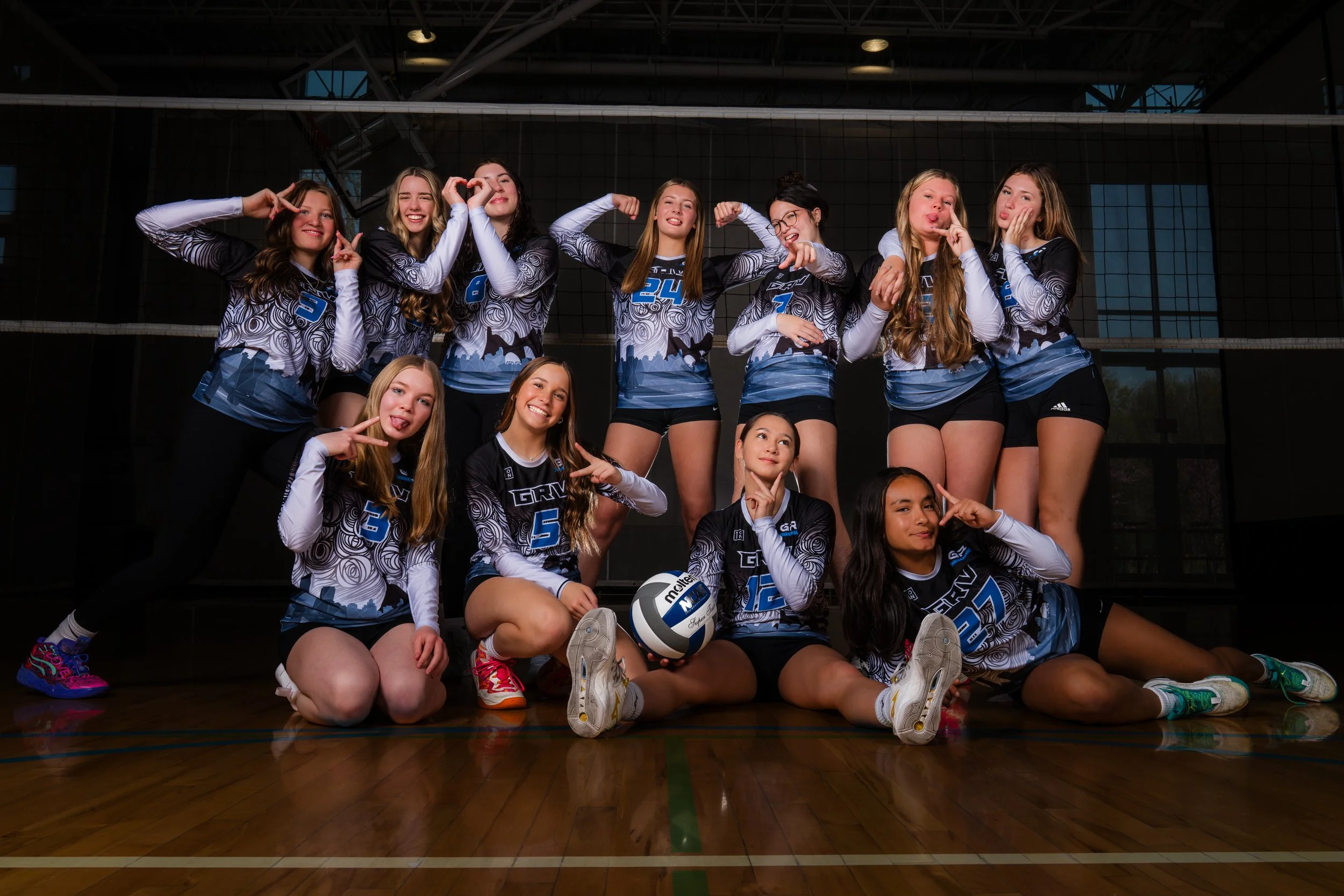 A volleyball team of twelve girls in matching jerseys posing inside a gymnasium. Some girls are standing, others kneeling or sitting, making playful gestures. The team is diverse in ethnicity and they are smiling and making peace signs or other fun g