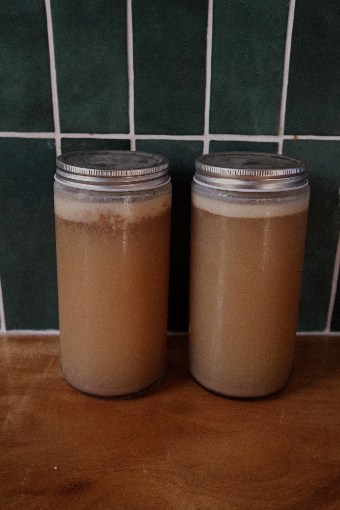Two glass jars with metal lids containing a light brown, creamy beverage, placed on a wooden surface against a green tiled wall.