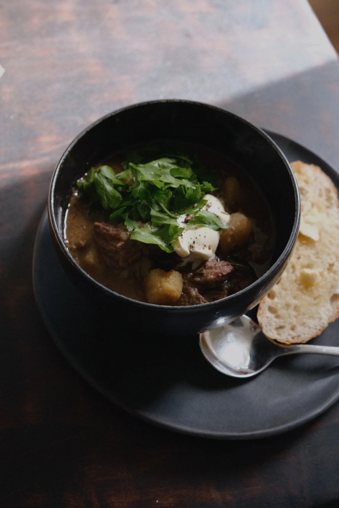 A bowl of beef stew topped with fresh herbs and sour cream, served with a slice of bread on a black plate with a spoon.