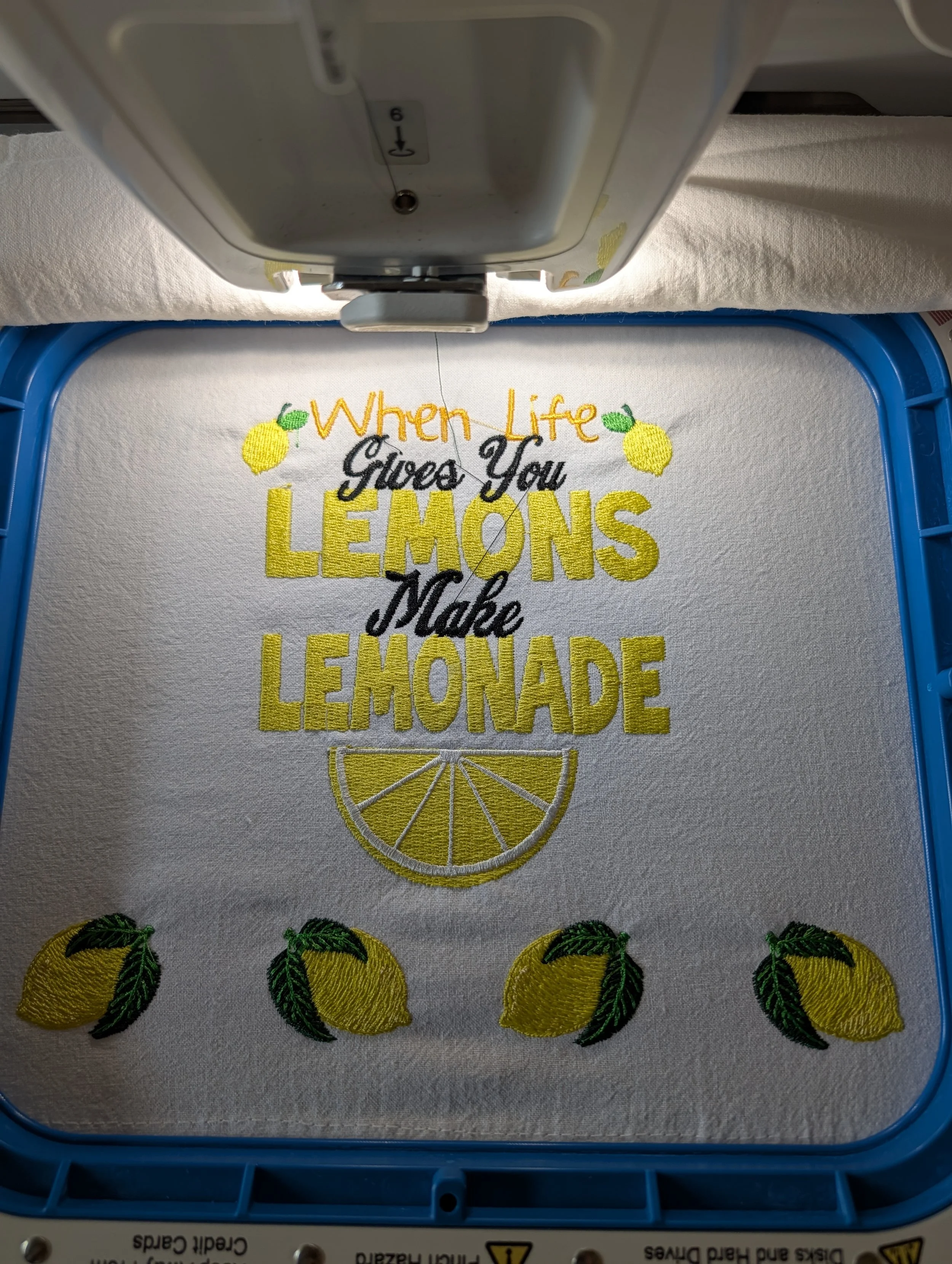 embroidery design that says when life gives you lemons make lemonade with a lemon slice underneath.  Stitched on a flour sack towel.