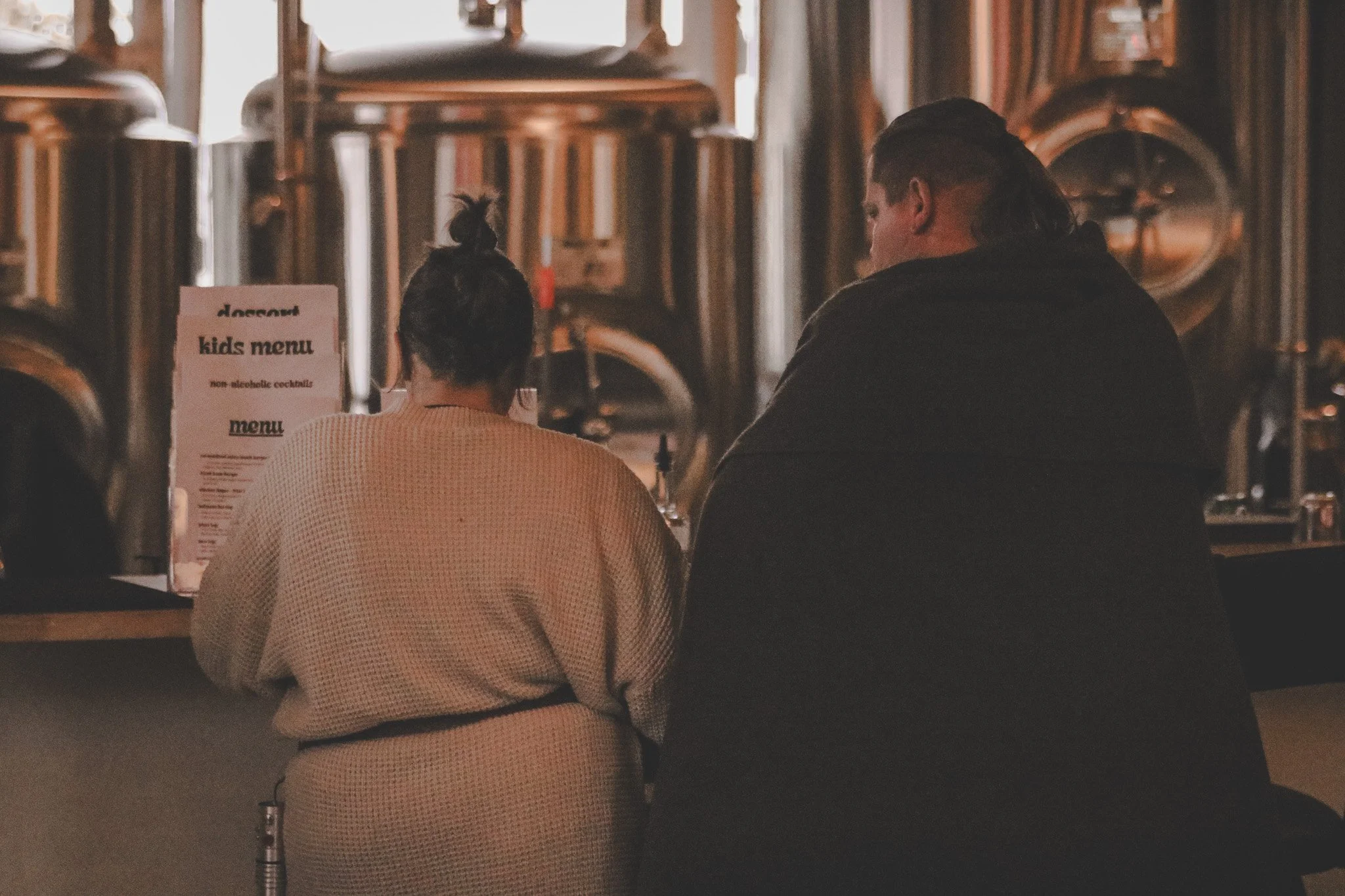 A woman with dark hair in a bun and a man with long hair sit at a bar or restaurant counter, with beer tanks in the background.