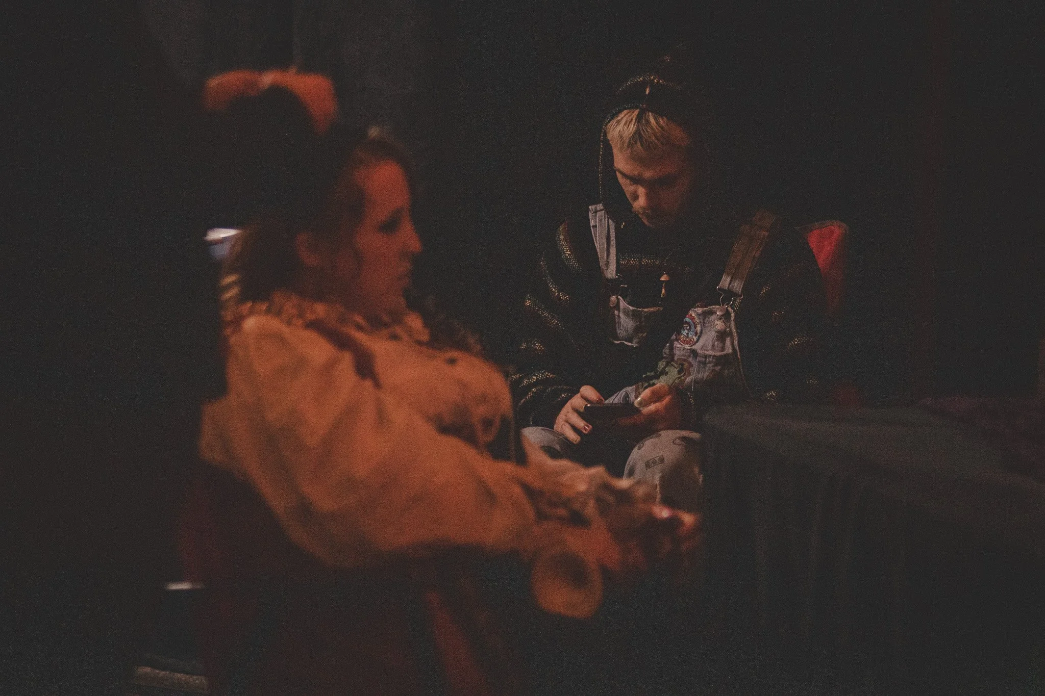 Two people sitting in a dark room, one woman with curly hair and a woman with short blonde hair, wearing a jacket with patches, looking at a device.