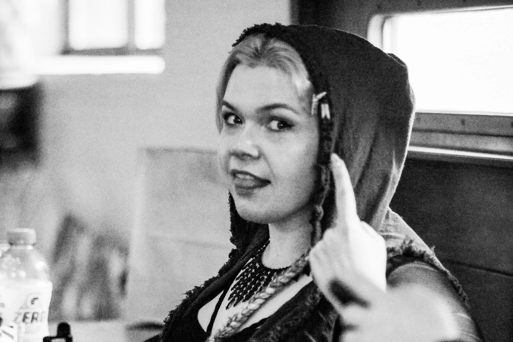 Black and white photo of a woman with braided hair wearing a hoodie, pointing to her head with her index finger, inside a vehicle.