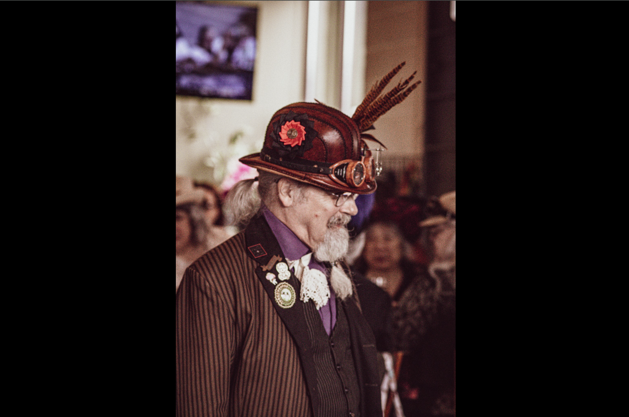 Man in a steampunk outfit with a decorated hat, including feathers and goggles, attending an event.