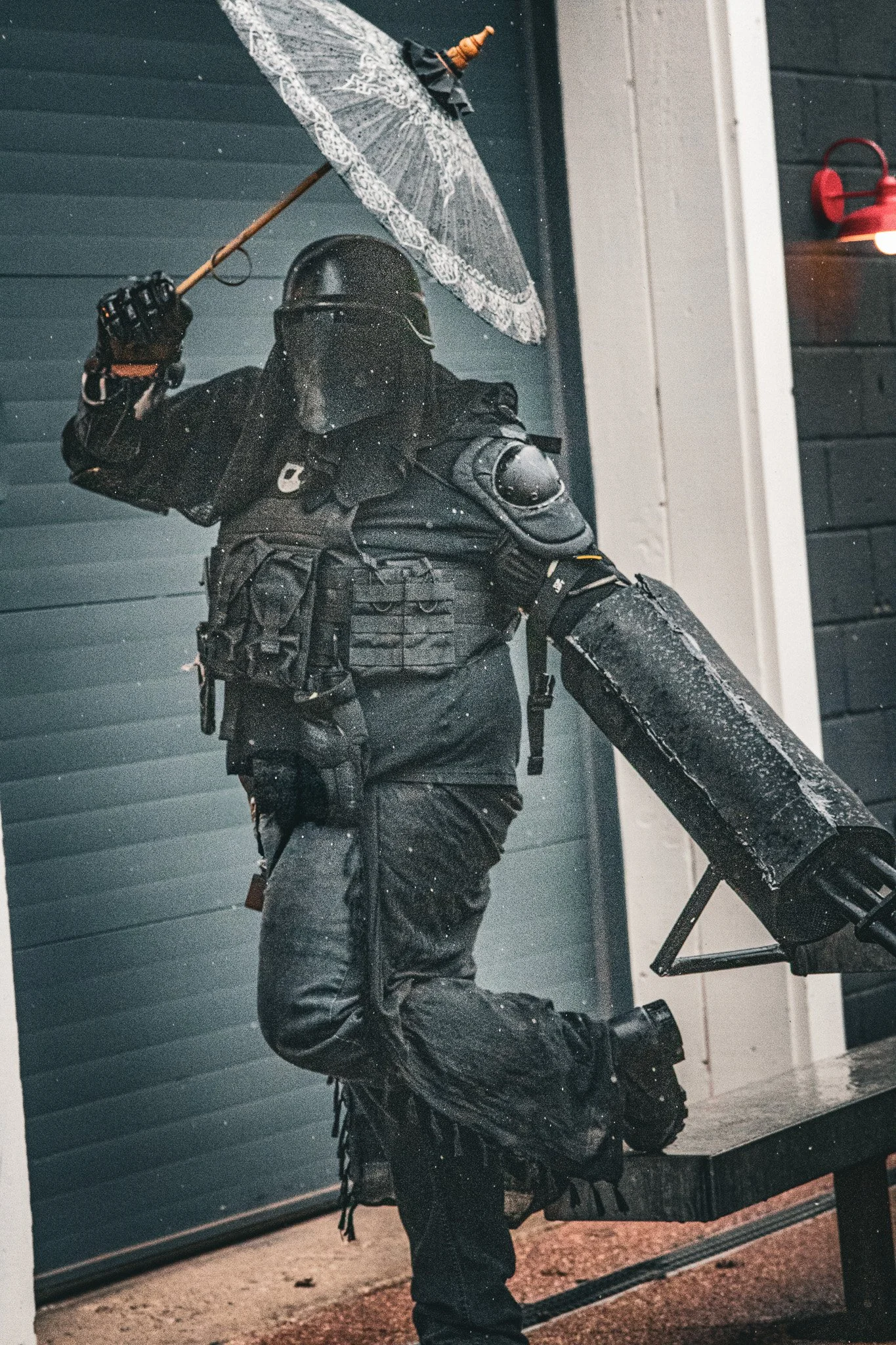 A person dressed in black tactical gear holding a lace umbrella standing outside at night, with a black shutter door and part of a house in the background.