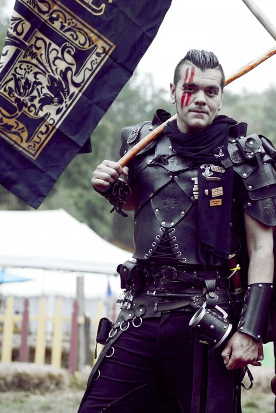 Person dressed in black leather armor with red face paint, holding a flag with a black and gold design, outdoors.