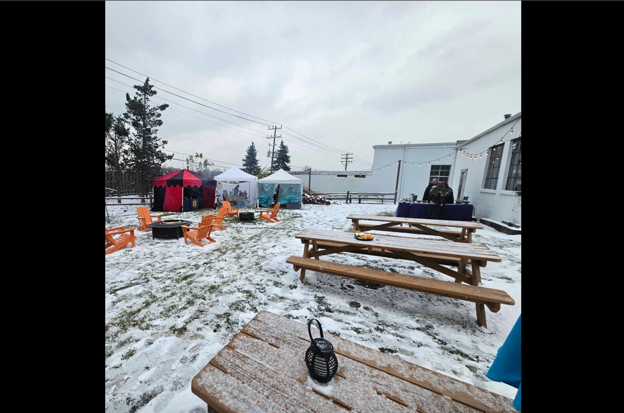 Snow-covered outdoor area with picnic tables and Adirondack chairs, several tents, a grill, and a building in the background, with trees and string lights visible.