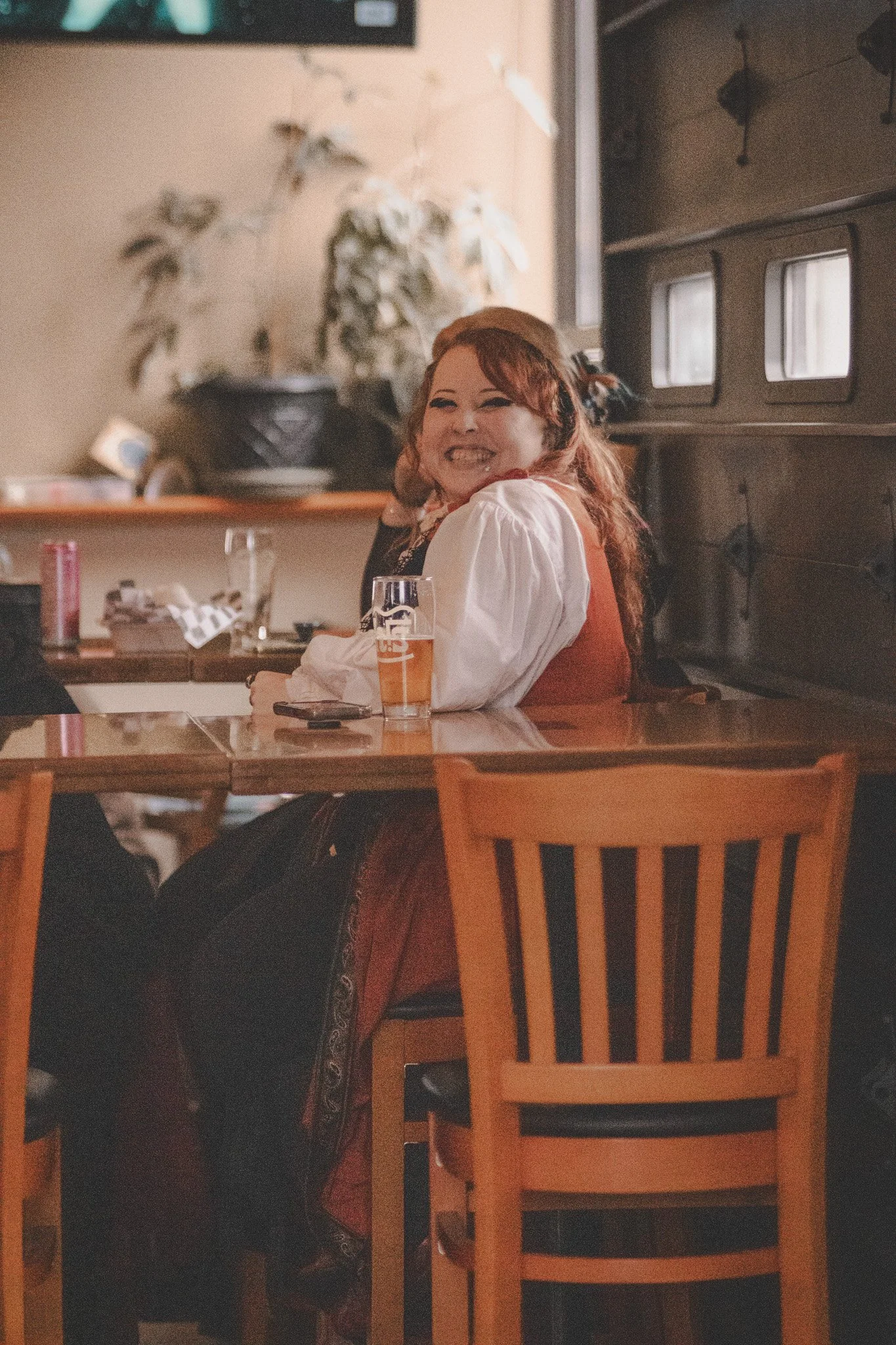 A woman with red hair and a white shirt smiles and laughs while sitting at a rustic wooden table in a cozy, warmly lit cafe or restaurant, with a pint of beer in front of her.