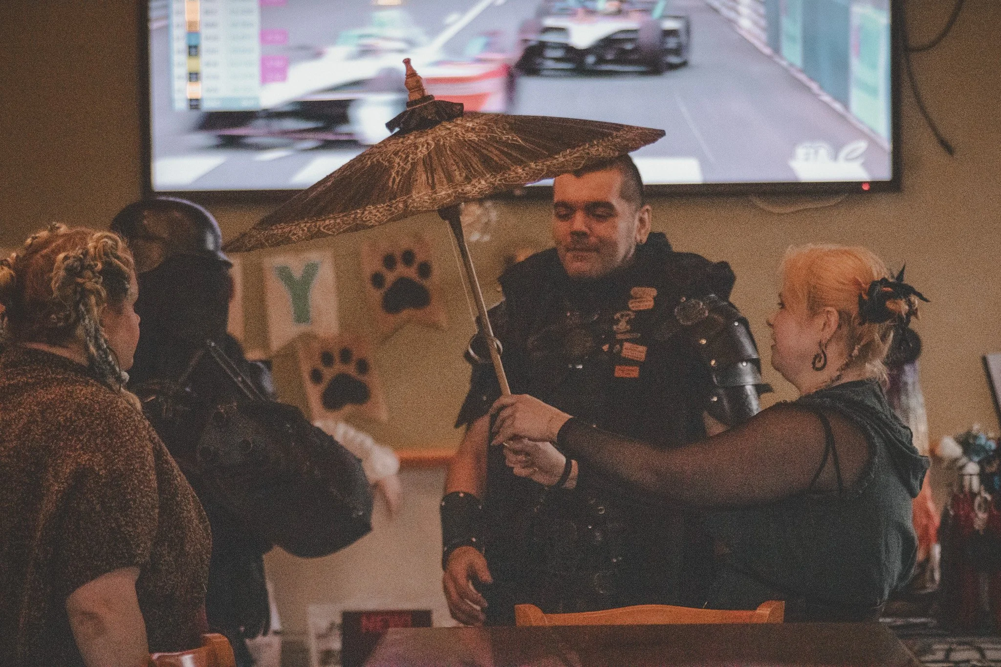 A man is holding a decorative umbrella over a woman in a dimly lit room decorated with dog-themed banners. Other people are present, some wearing black clothing, with a TV screen showing a racing event in the background.
