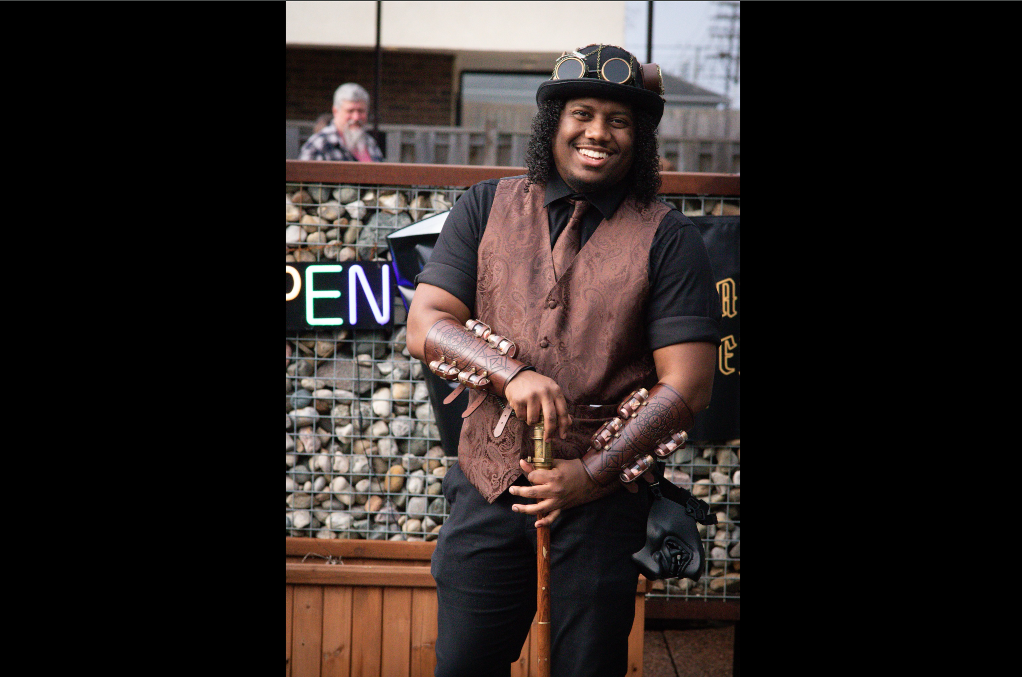 Person in steampunk outfit with brown vest, goggles on hat, arm bracers, and cane, standing in front of rock-filled fence and neon sign.