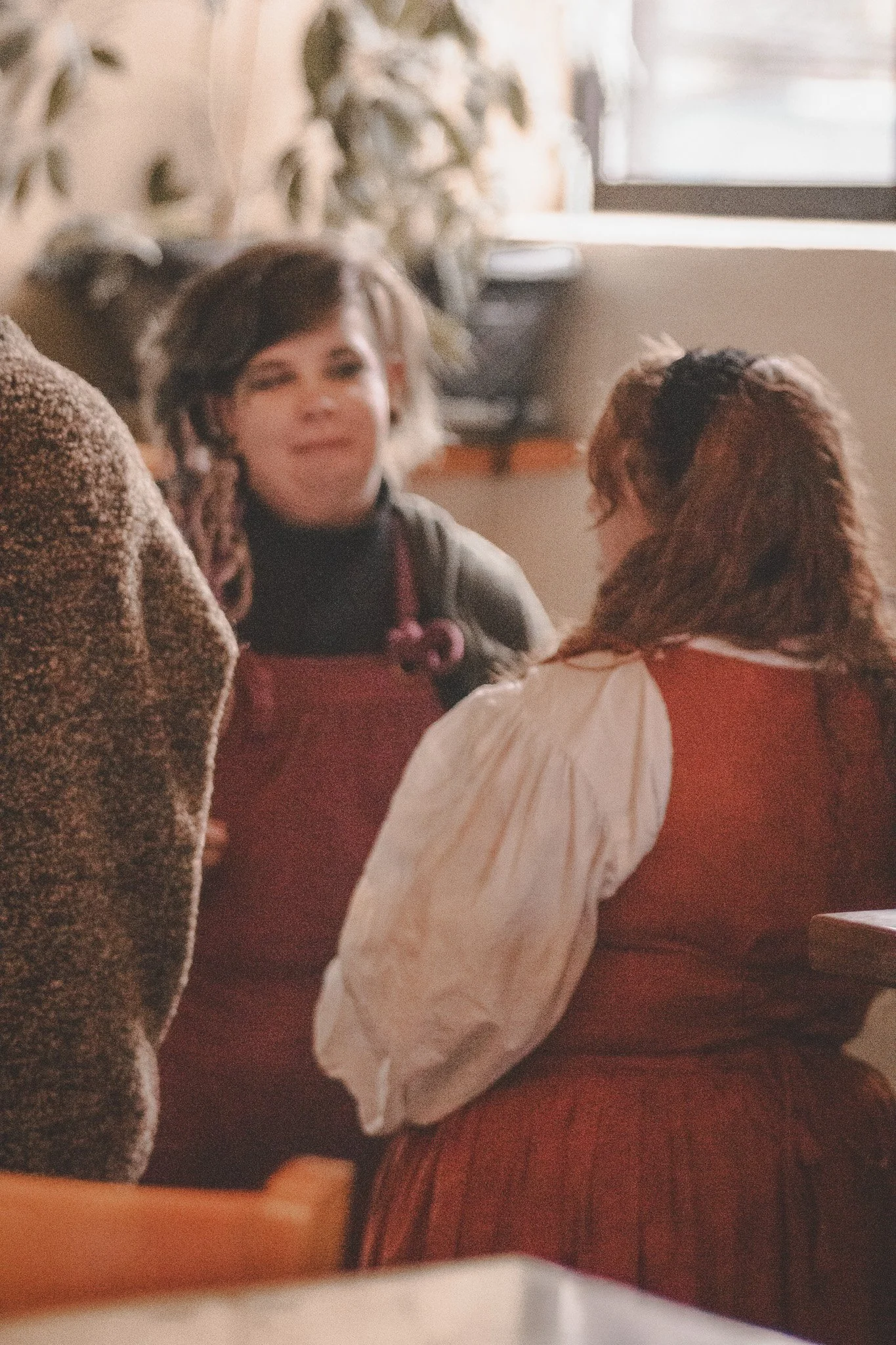 Two women having a conversation inside a cafe, with plants and a window in the background.