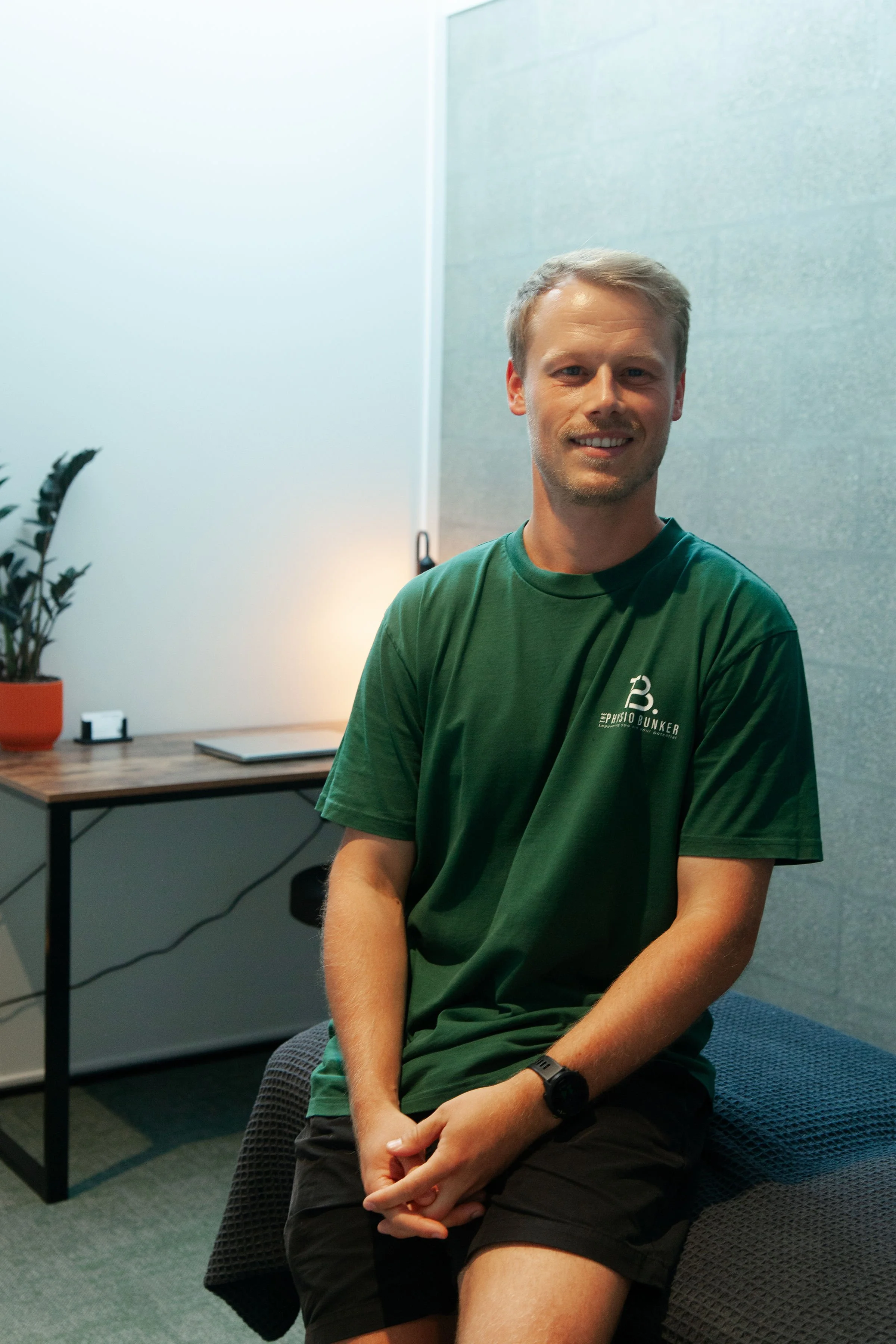 A young man with short blond hair, wearing a green T-shirt with the logo 'The Physio Bunker,' is sitting on a gray cushioned chair in an office or clinic environment, smiling. There is a wooden desk with a plant, a small black and white object, a closed book, and a laptop in the background, along with a gray wall and some lighting.