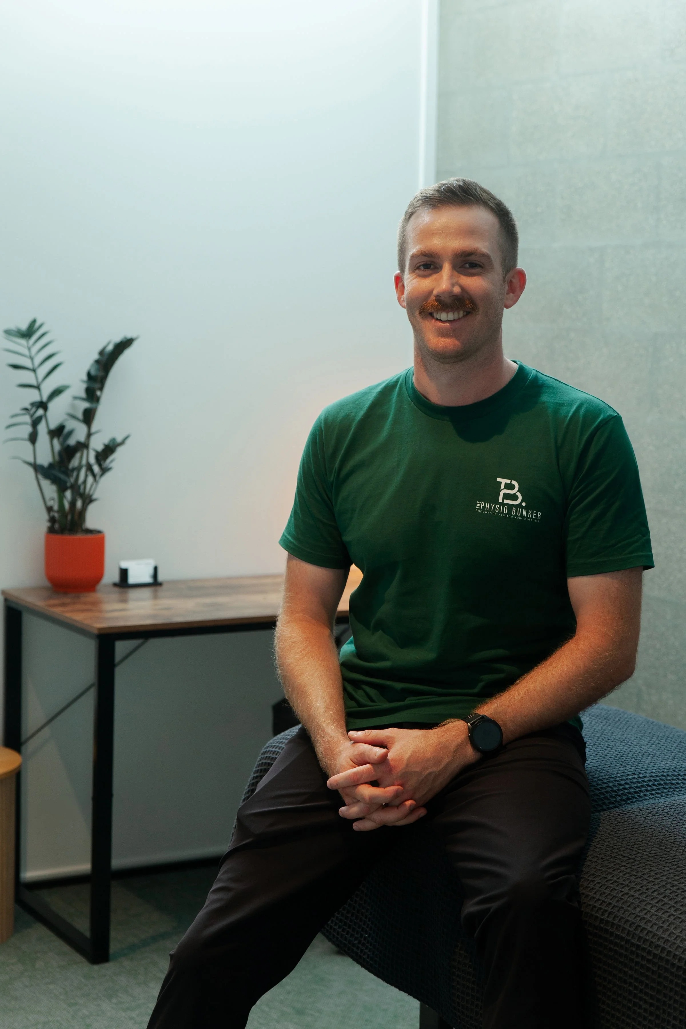 A smiling man with a mustache and short brown hair, wearing a green T-shirt with 'Physio Bunker' logo, sitting on a chair in an office with gray walls, a wooden table, a potted plant, and a small electronic device in the background.