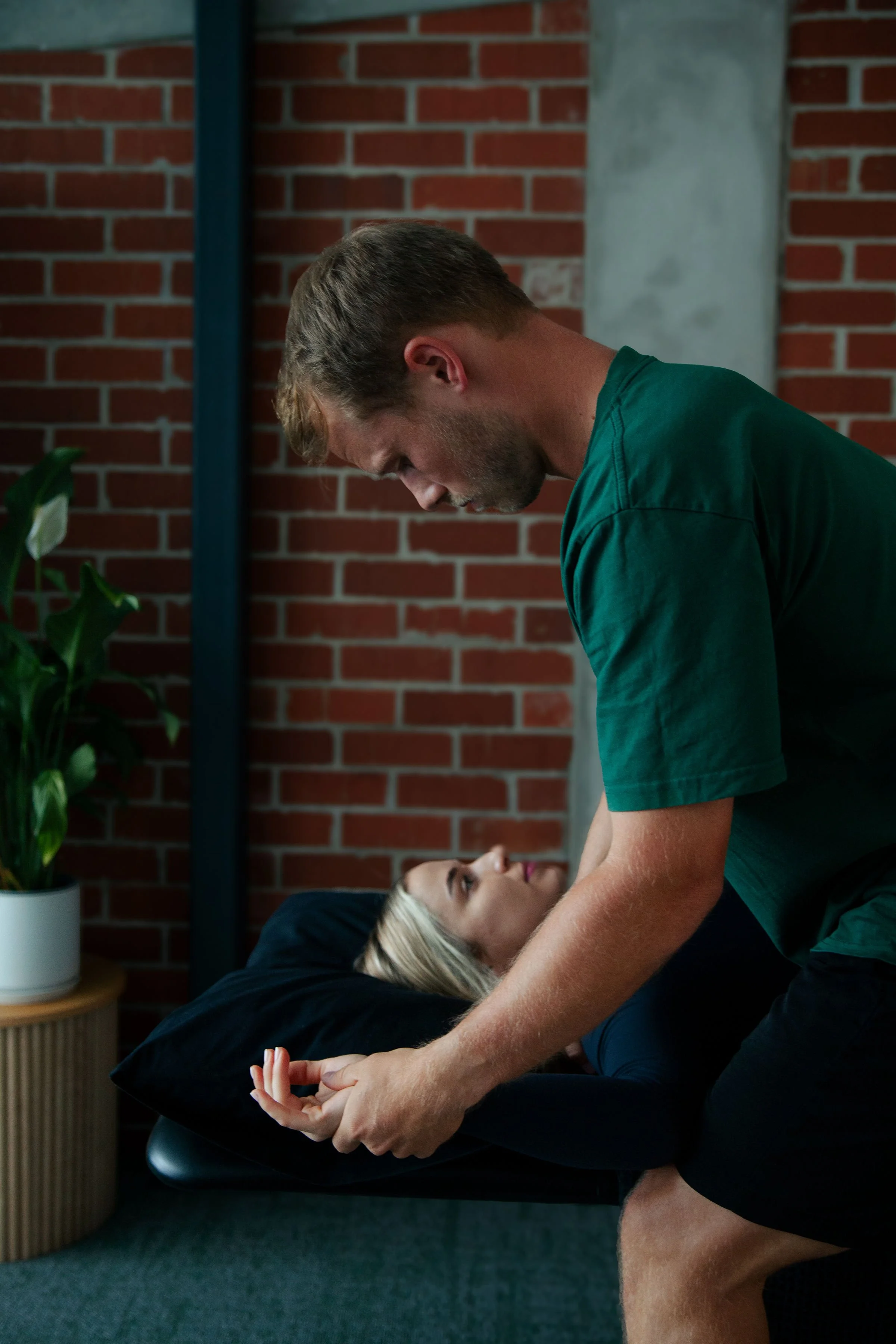 A man with short brown hair and a beard provides chiropractic or massage therapy to a woman lying on a treatment table. The woman has blonde hair and is looking up at the man while he works on her. The setting has a brick wall, a potted plant, and a small side table.
