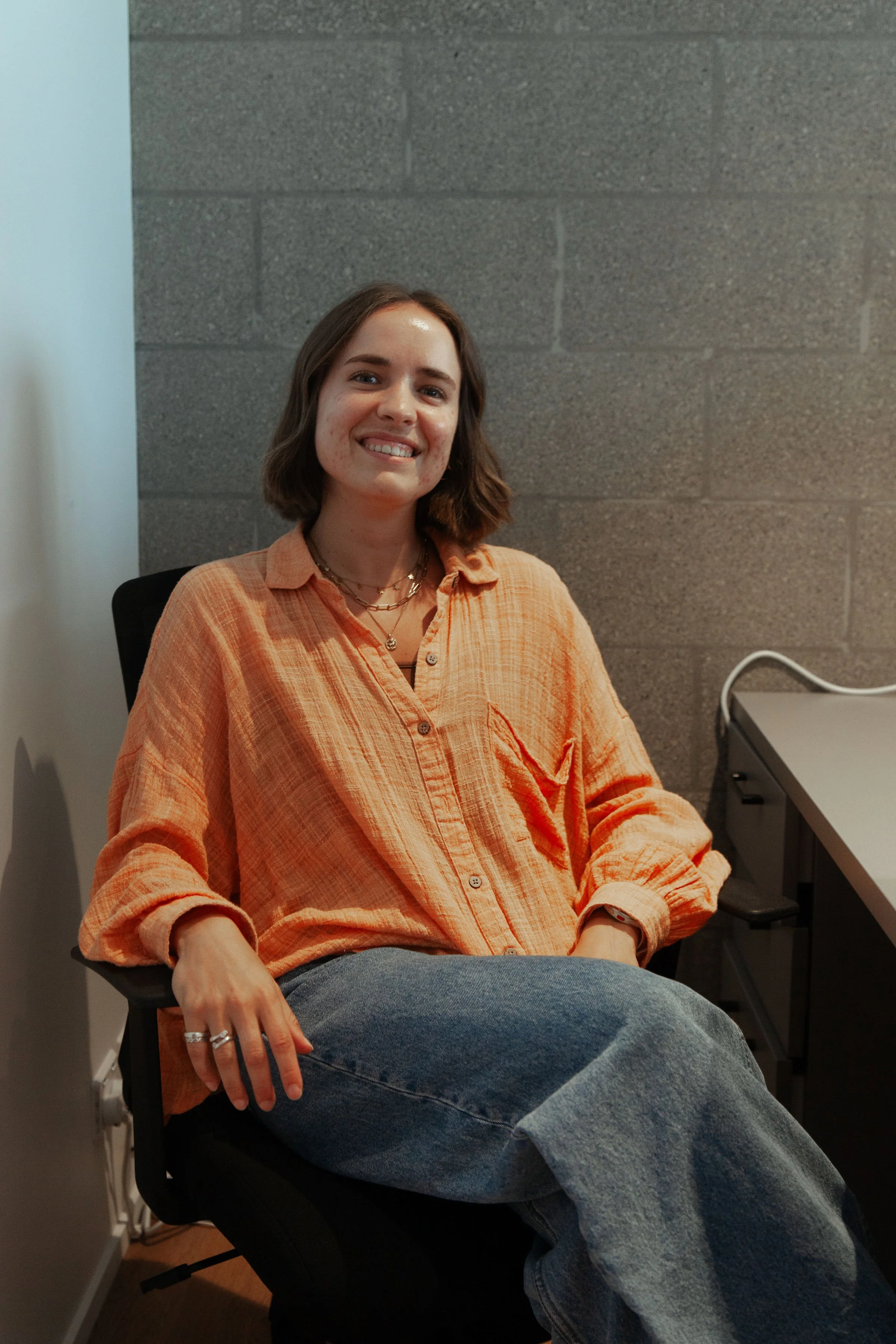 A young woman with short brown hair sitting in a black chair, smiling, wearing an orange button-up shirt and blue jeans against a gray concrete block wall.