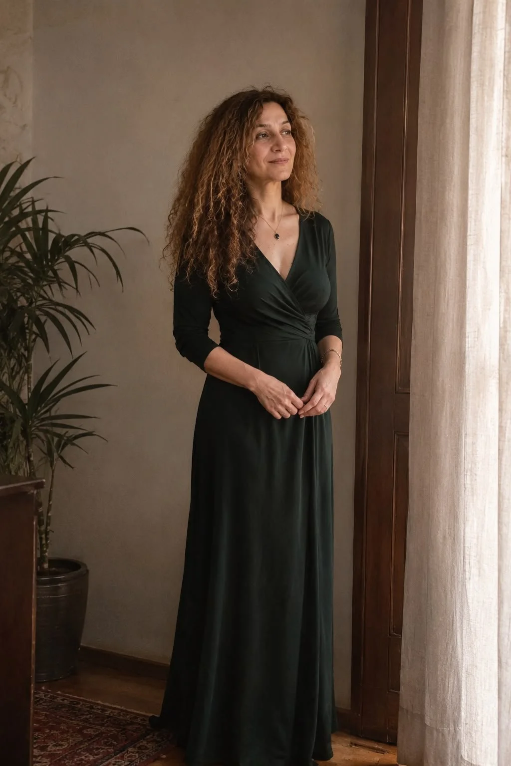 A woman with curly brown hair in a black dress standing near a window with beige curtains, indoors with a potted plant nearby.