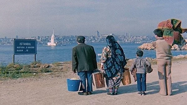 A family of four, including an elderly man, woman, and two children, standing on a shoreline with a view of Istanbul across the water, with some carrying bags and one person holding an umbrella.