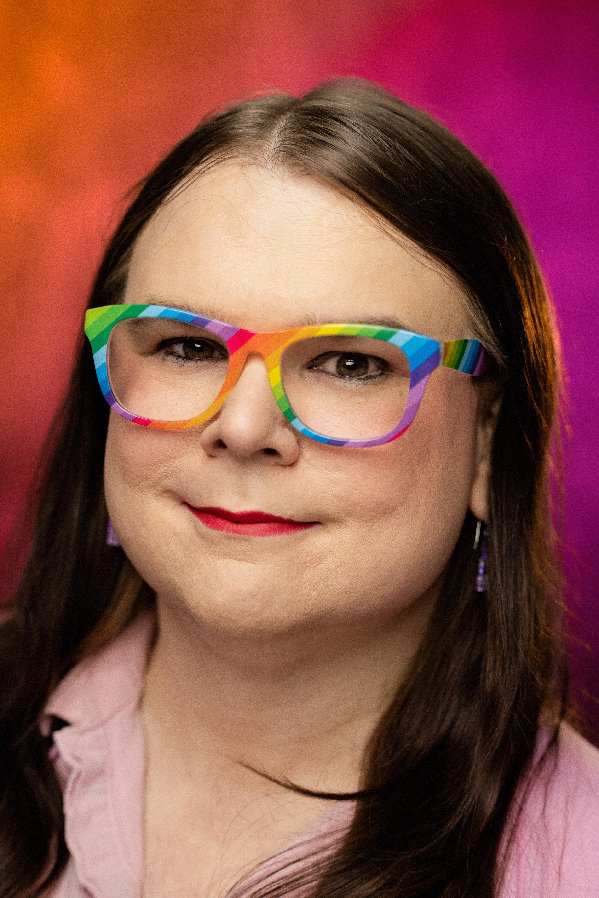 Close-up portrait of a woman with long brown hair, wearing black glasses, and smiling softly. She has light skin and is dressed in a dark top against a light background.