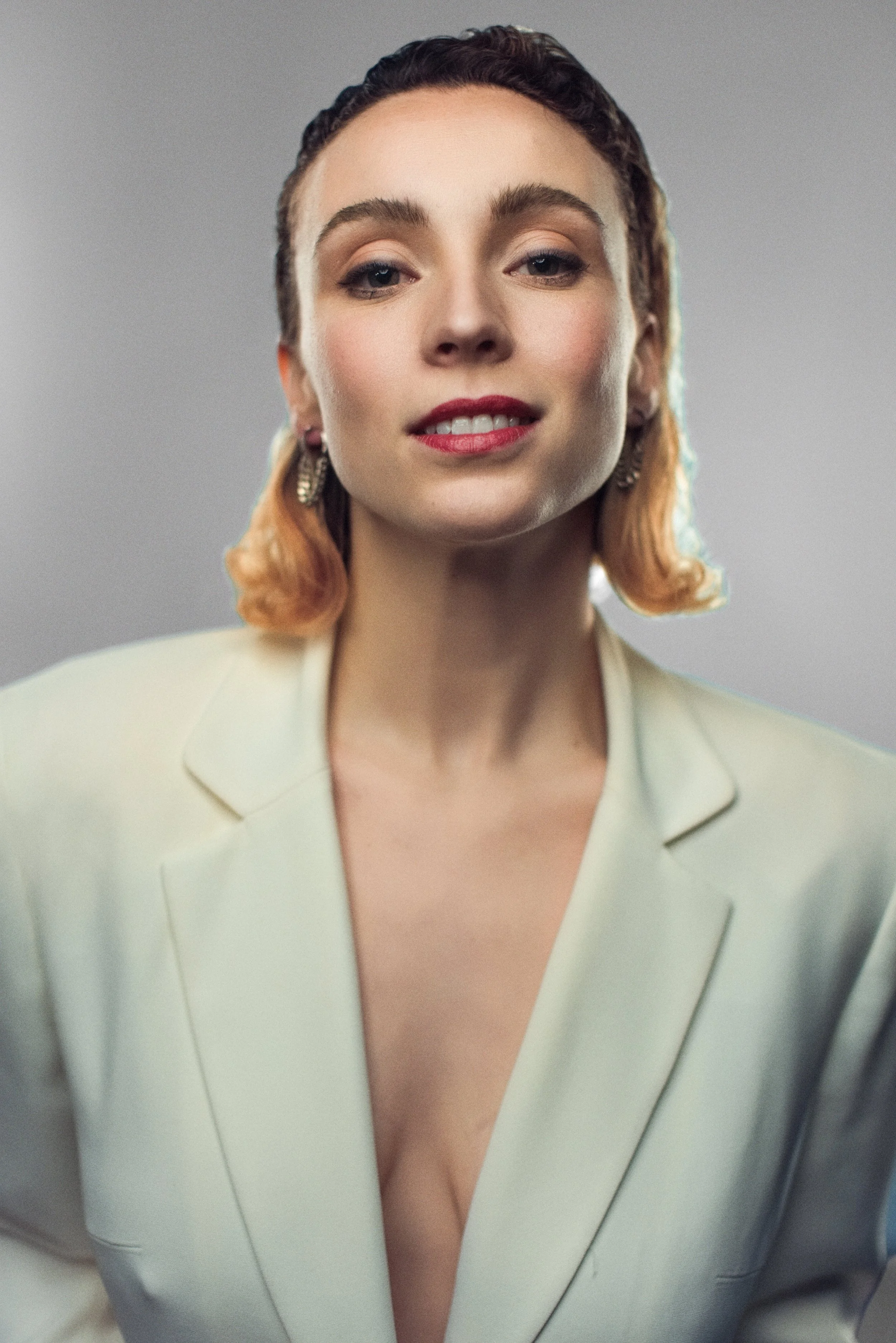 A young woman with curly blonde hair smiling, wearing a maroon tank top, against a vibrant orange background.