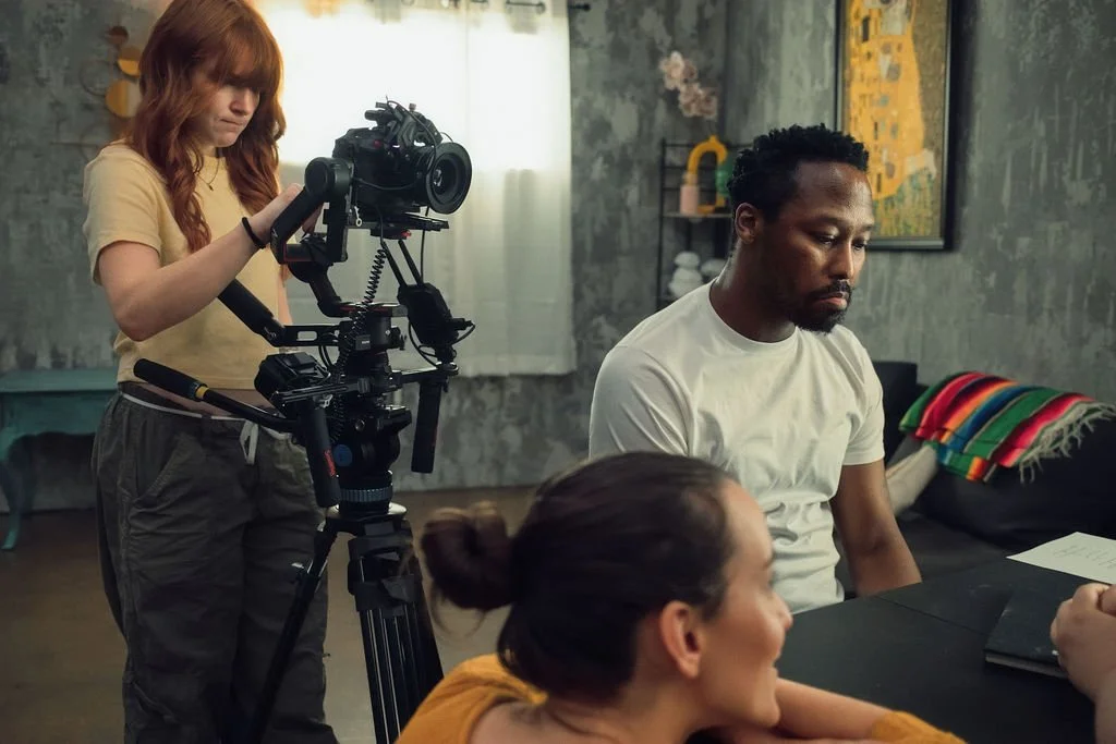 A woman operating a professional film camera on a tripod while two people sit at a table, one man and one woman, engaged in a discussion in a cozy room.
