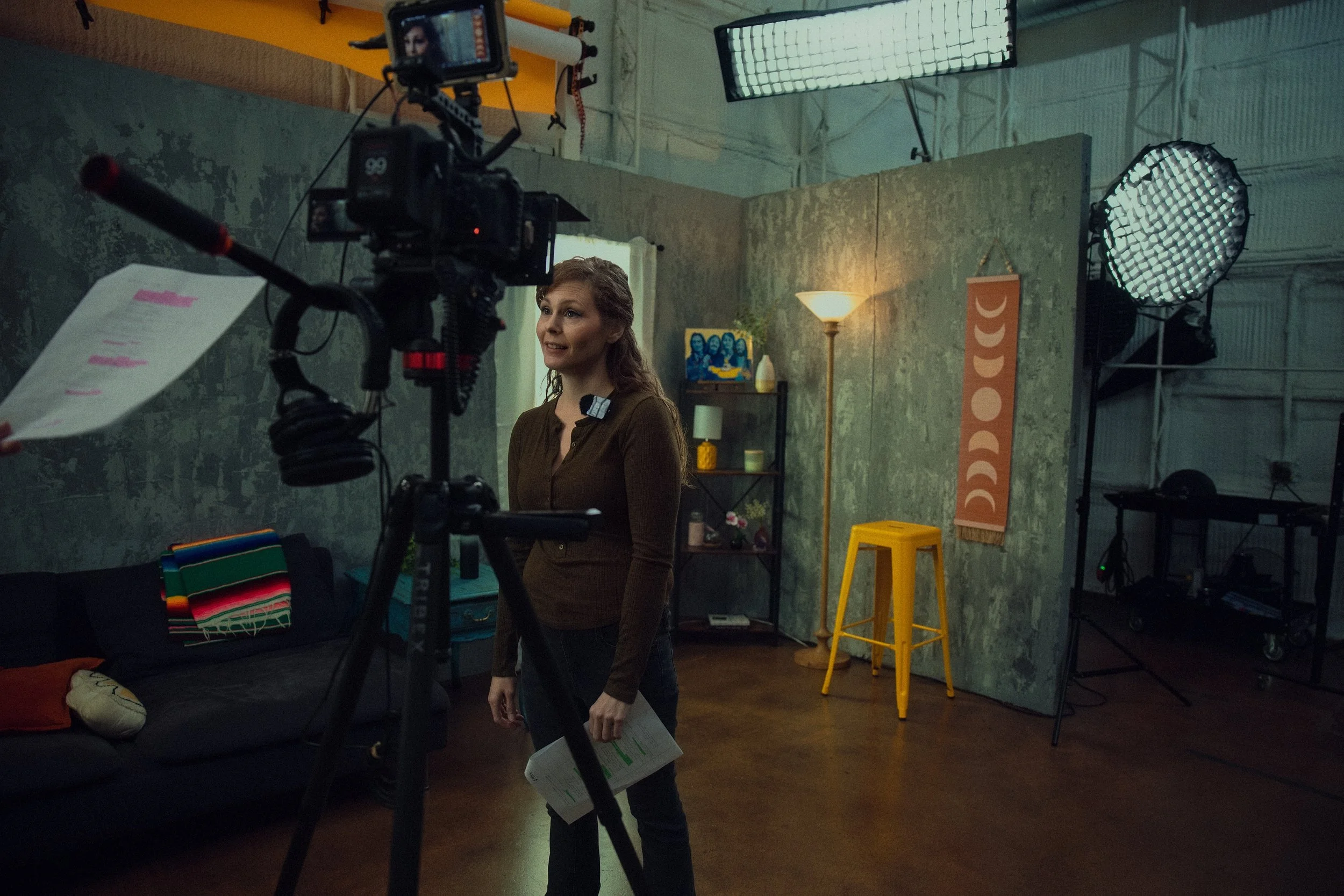 Woman standing in a recording studio or set with professional lighting and camera equipment, holding papers, with a sofa and decorative items in the background.